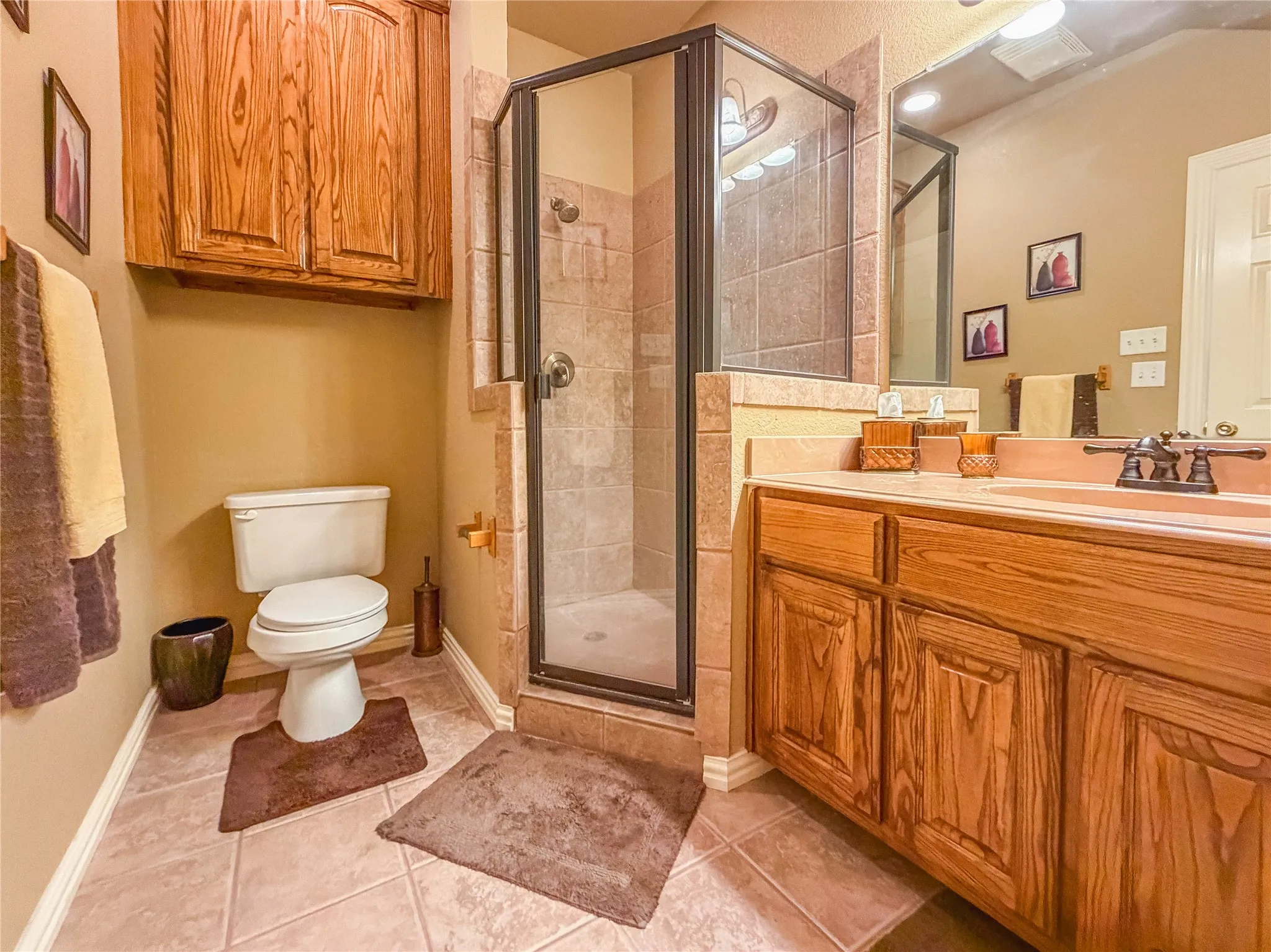 Tiled flooring with an enclosed shower, wood-finish vanity with an integrated sink, and an overhead wood-finish cabinet