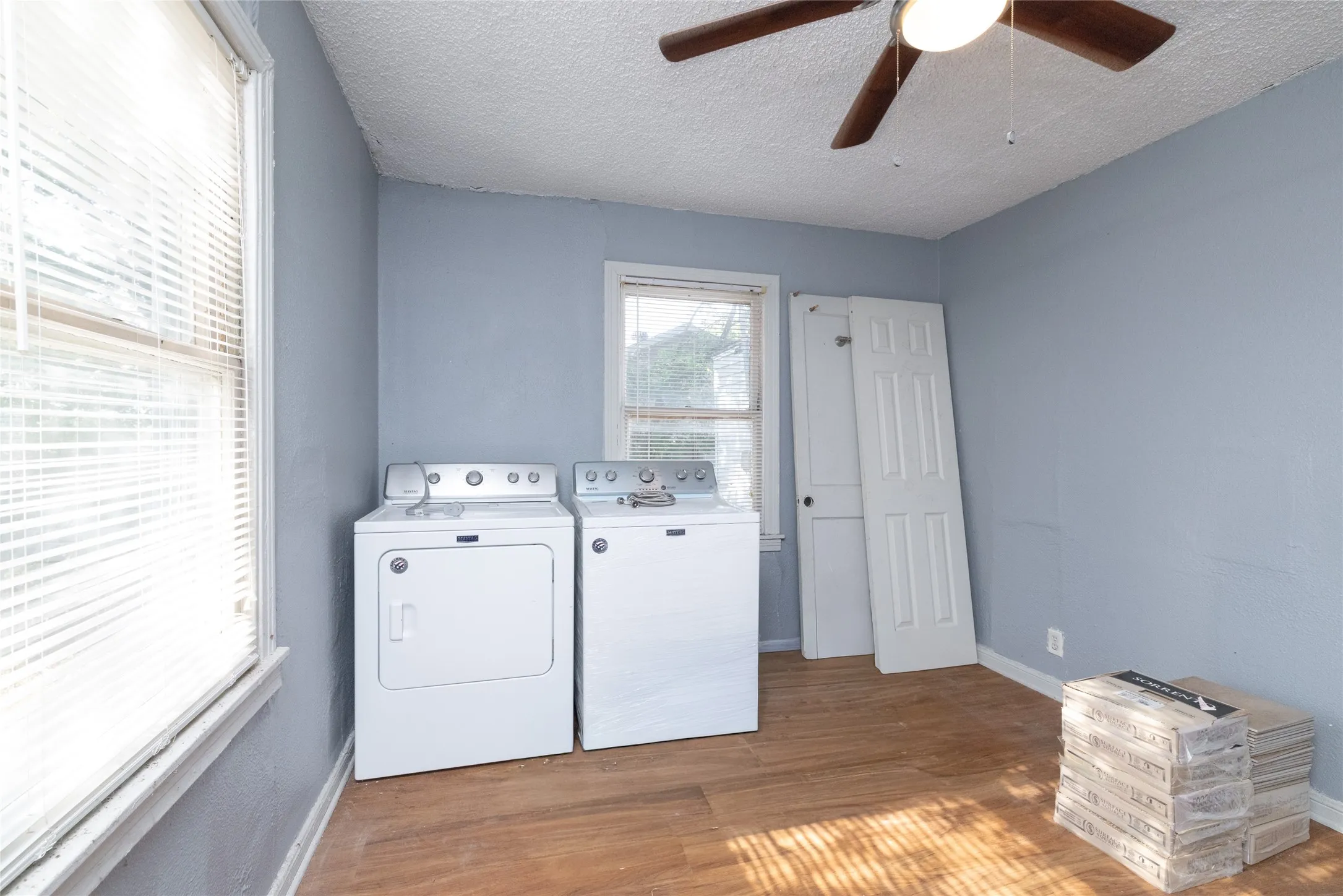 Wood-finish flooring, ceiling fan with integrated lighting, two windows with blinds, light blue wall paint, and white trim