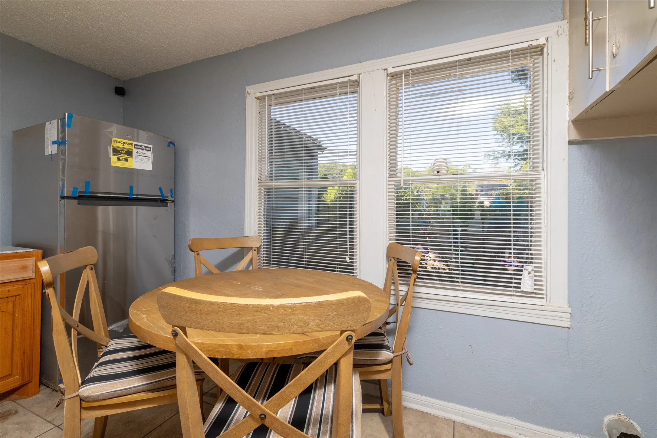 Two windows with blinds, light blue wall paint, light wood-finish cabinetry, and a stainless steel refrigerator
