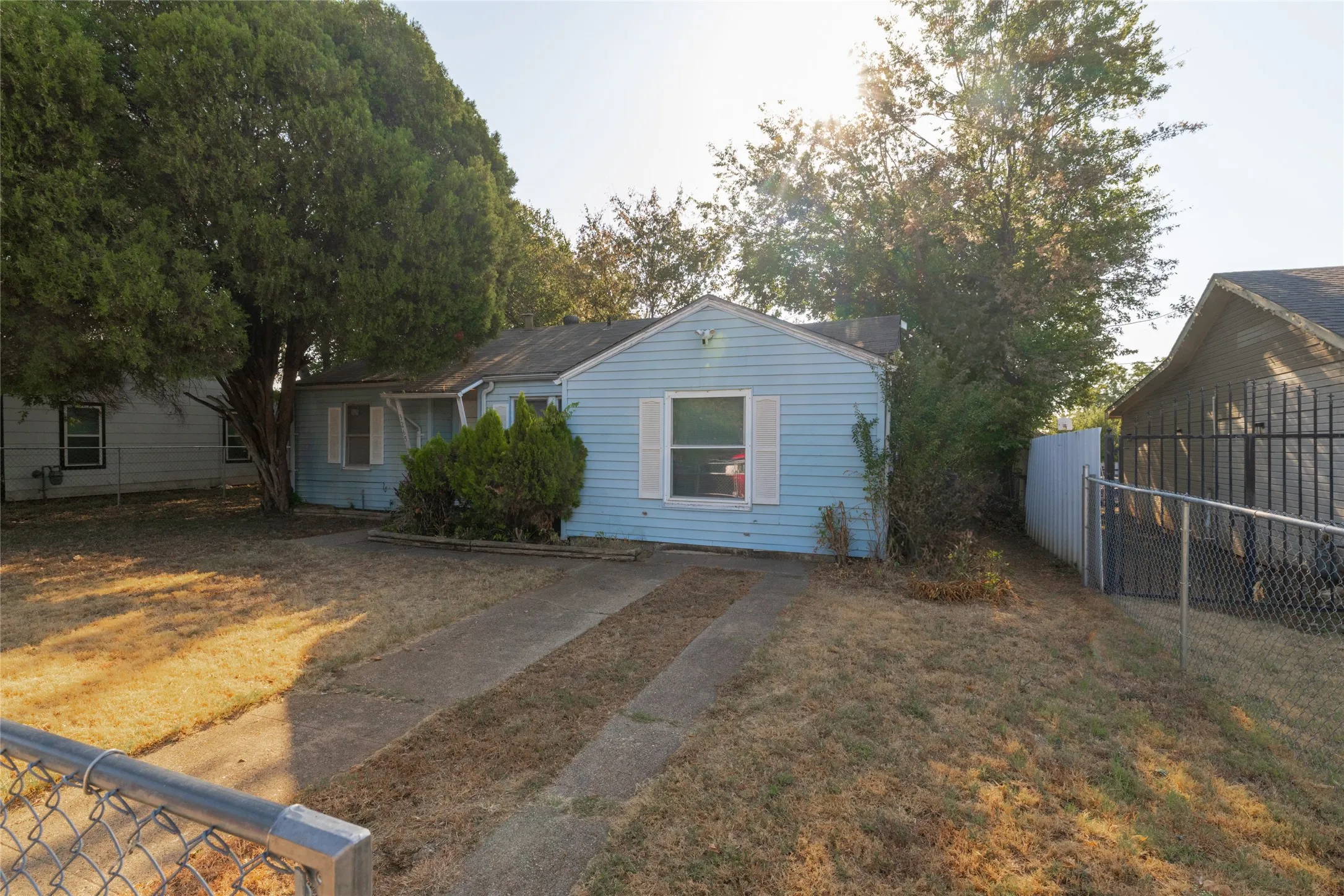 Exterior featuring light blue siding, white window shutters, mature trees, and a concrete driveway