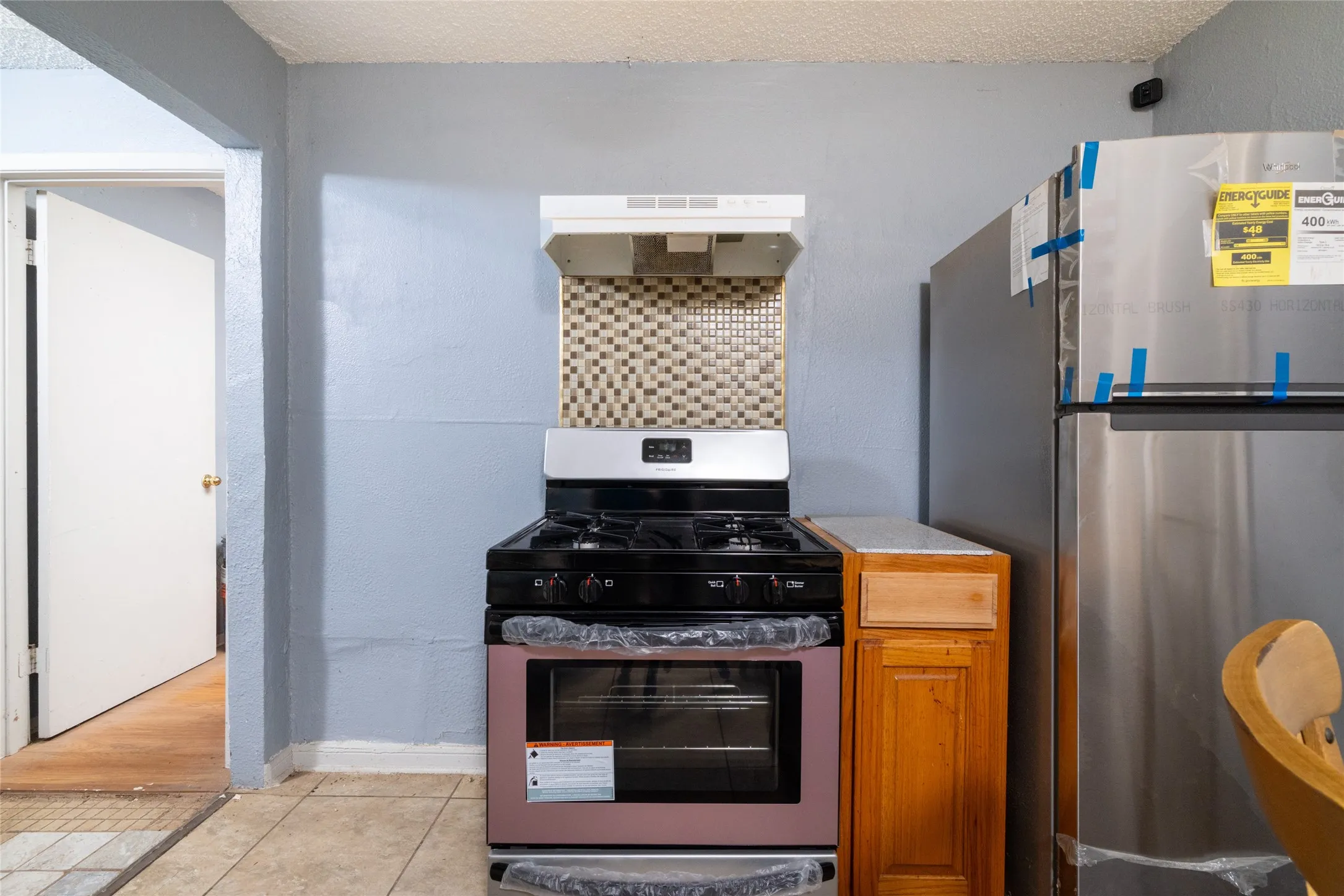 Stainless steel refrigerator, gas range with oven, mosaic tile backsplash, range hood, and wood-finish cabinetry