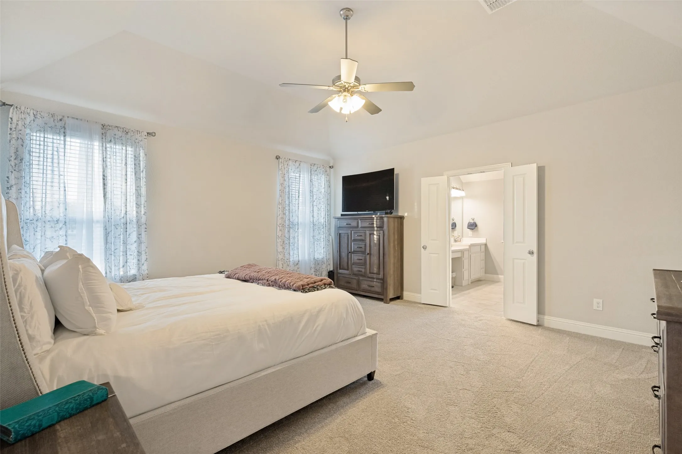 Vaulted ceiling with ceiling fan, dual windows, carpeted flooring, and a double-door entryway to a bathroom with dual vanities