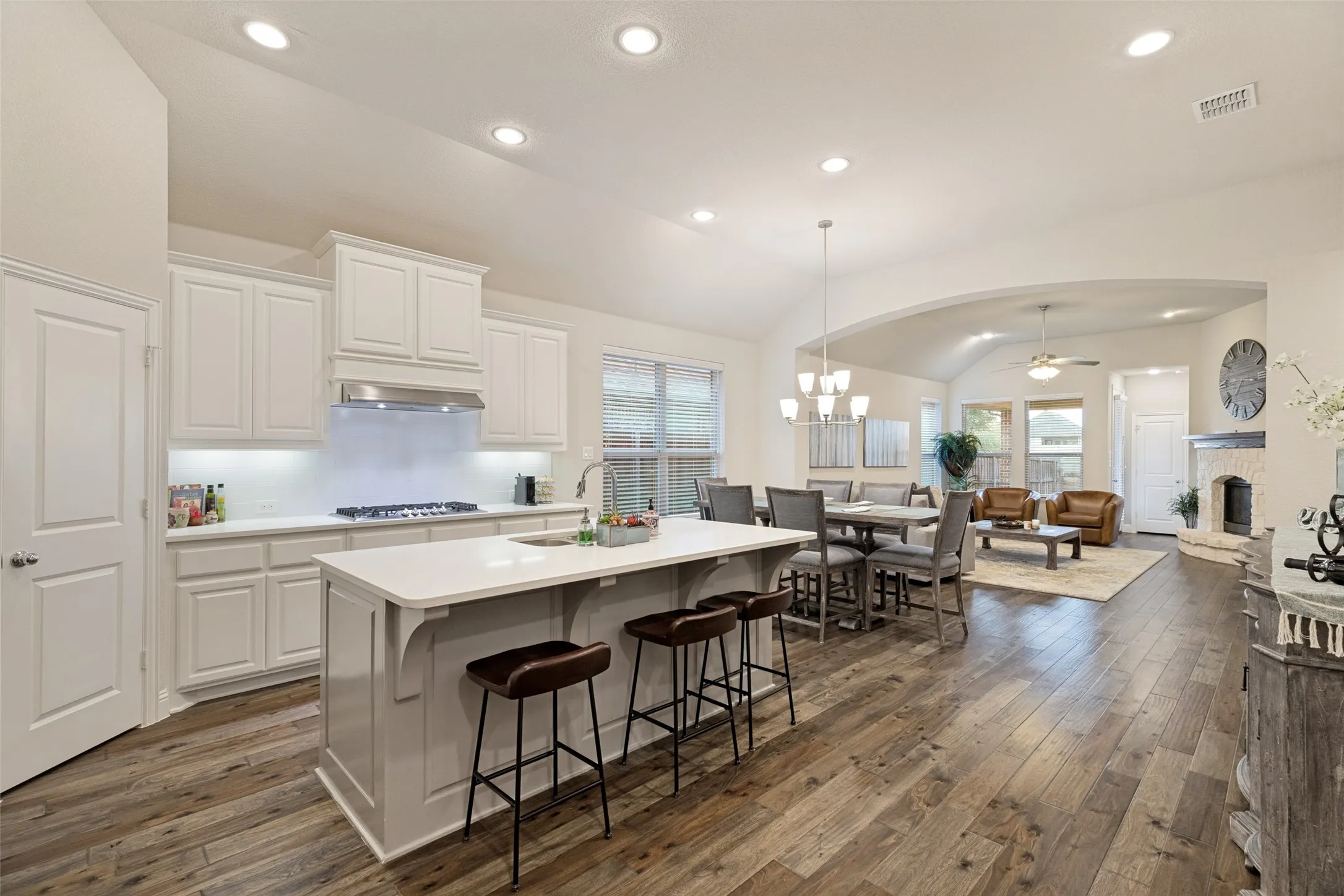 Open-concept living space with wood-finish flooring, large kitchen island, white cabinetry, a built-in fireplace, and vaulted ceilings