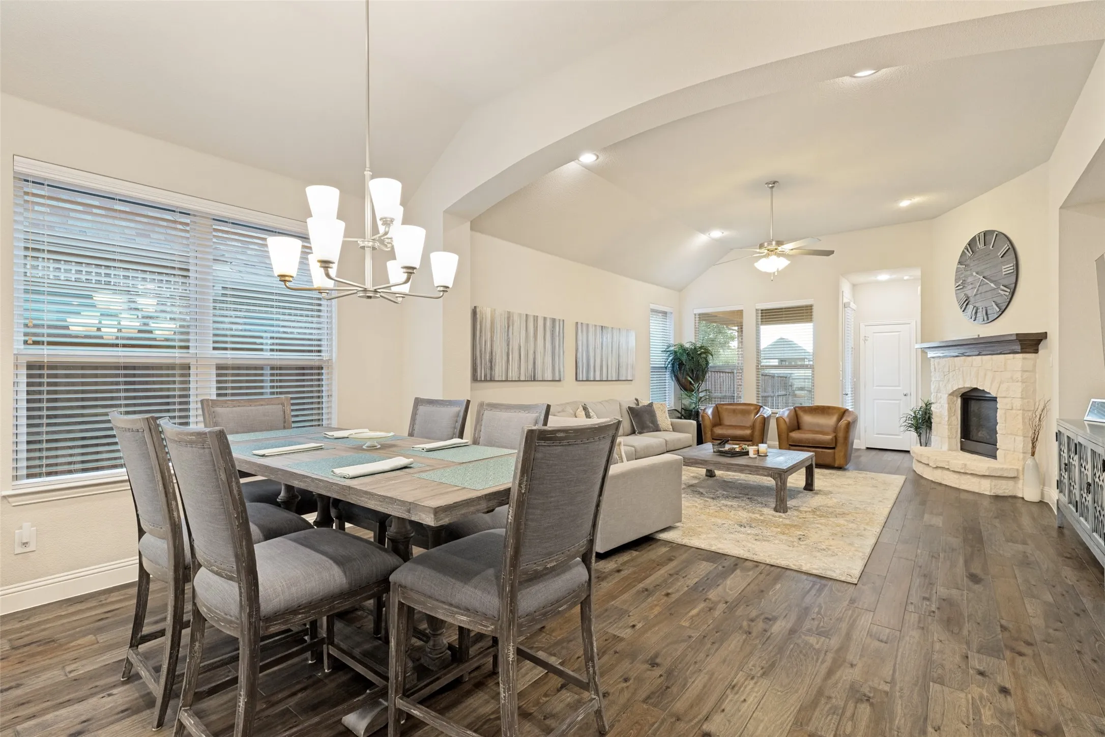 Vaulted ceilings featuring recessed lighting, a stone-faced fireplace with a wood mantle, wood-finish flooring, and multiple windows with blinds