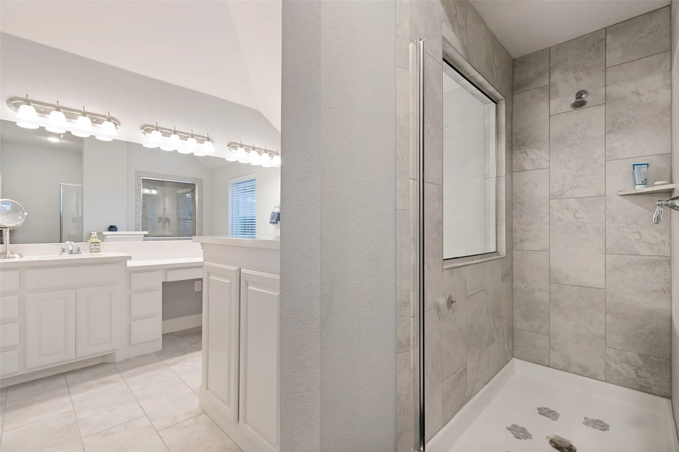 White vanity cabinetry, dual vanity sinks, extensive vanity mirror, recessed dressing table, and a glass-enclosed shower with tile surround