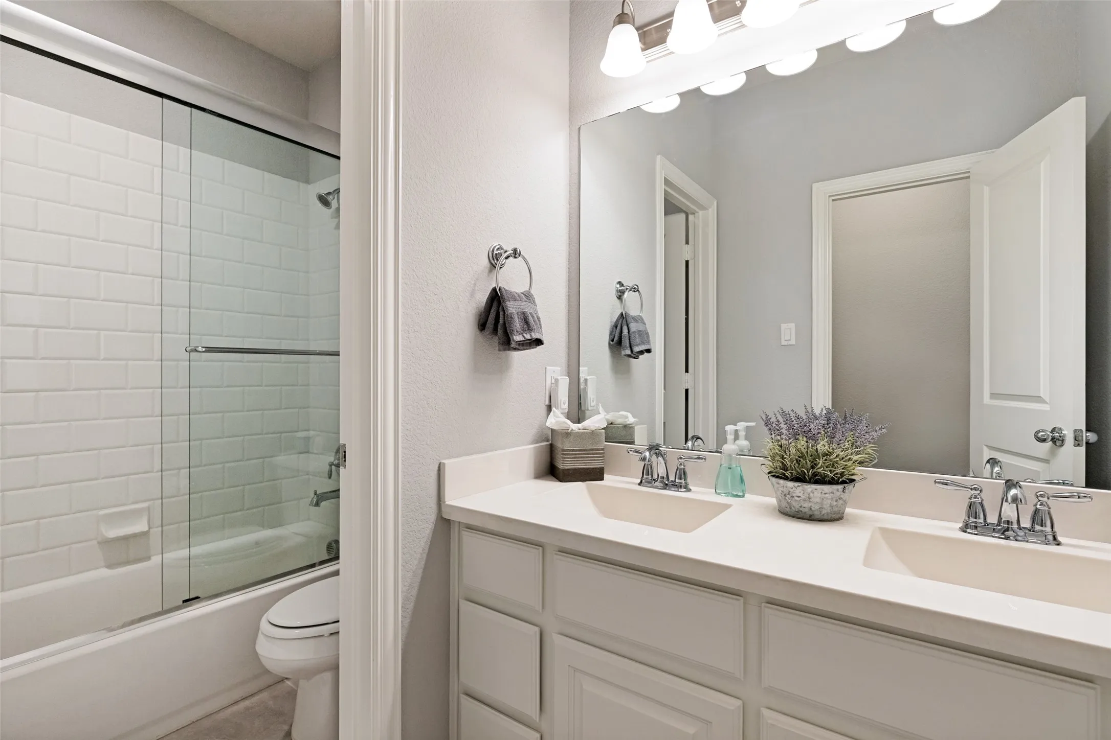 Dual vanity with integrated sinks, full-width mirror, white cabinetry, and a tub-shower combination with subway tile surround