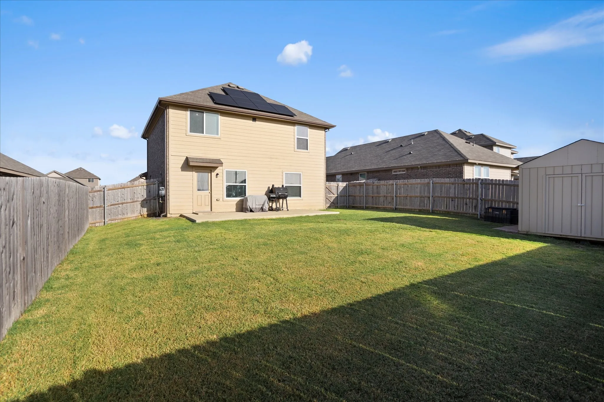 Expansive fenced backyard with a patio, featuring a storage shed and solar panels on the roof