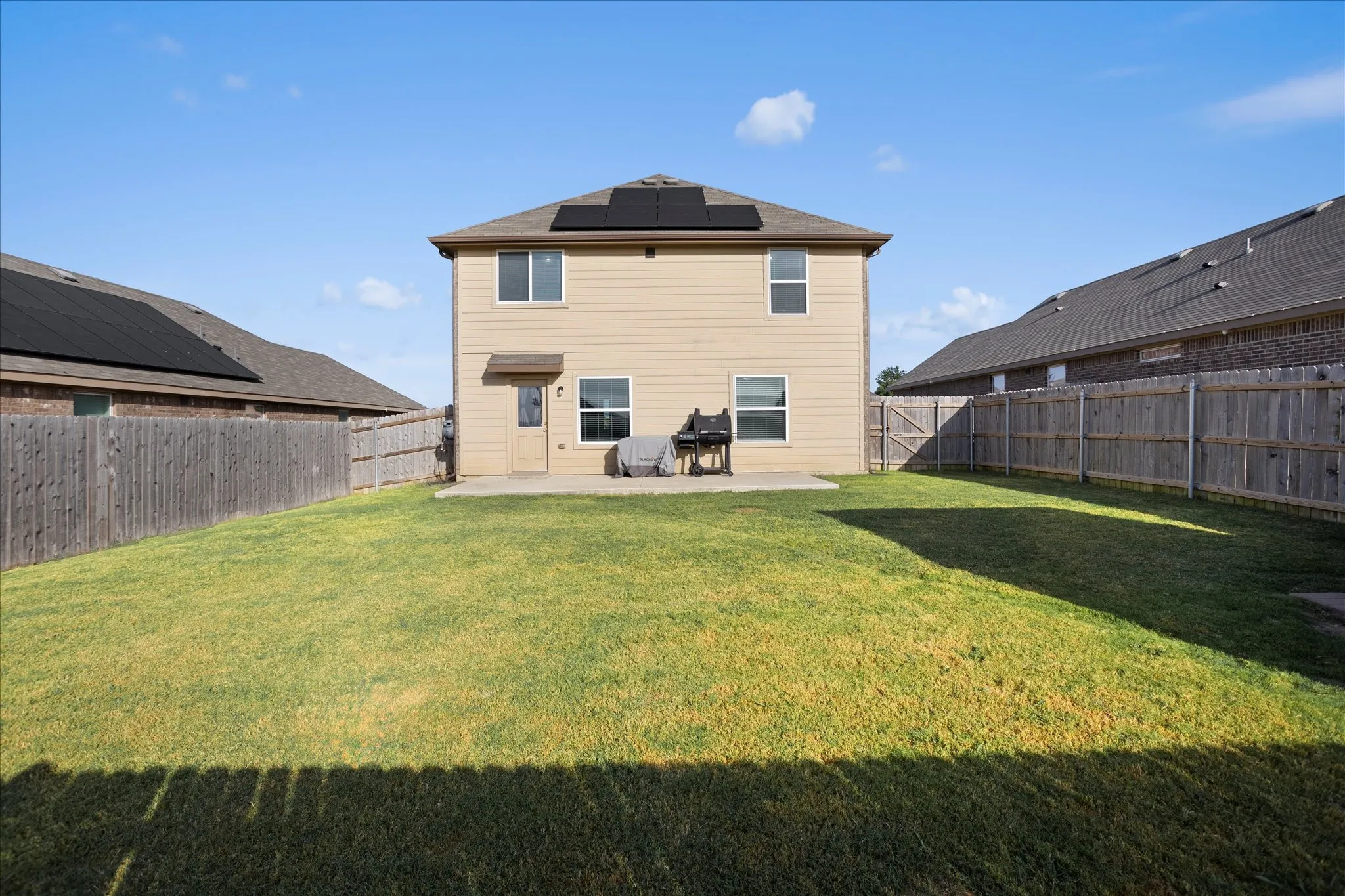 Expansive grassy backyard featuring a full privacy fence, concrete patio, and solar panels on the roof