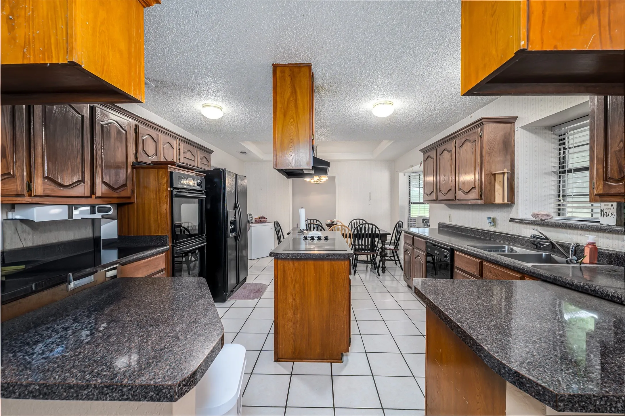 Kitchen featuring wood cabinetry, dark granite-style countertops, white tile flooring, and integrated black appliances