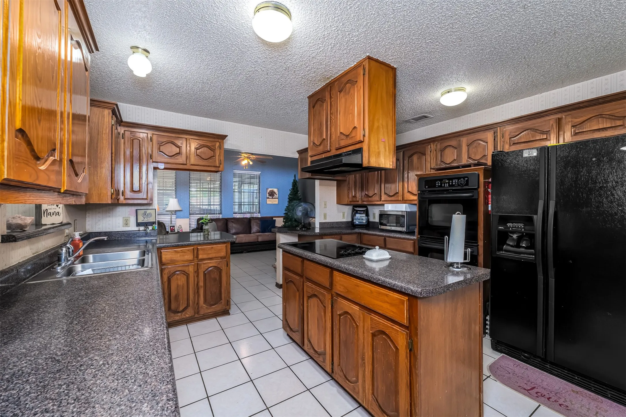 Kitchen featuring wood cabinetry, an island with a built-in cooktop, dark speckled countertops, and white square tile flooring
