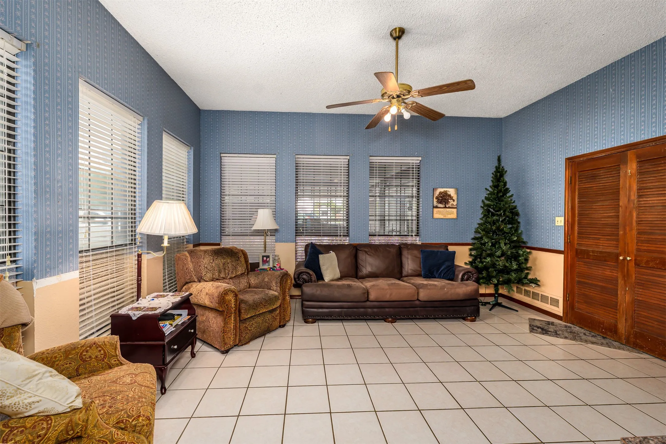 Spacious living area featuring tile flooring, multiple windows with blinds, a ceiling fan, and built-in wood-finish louvered doors