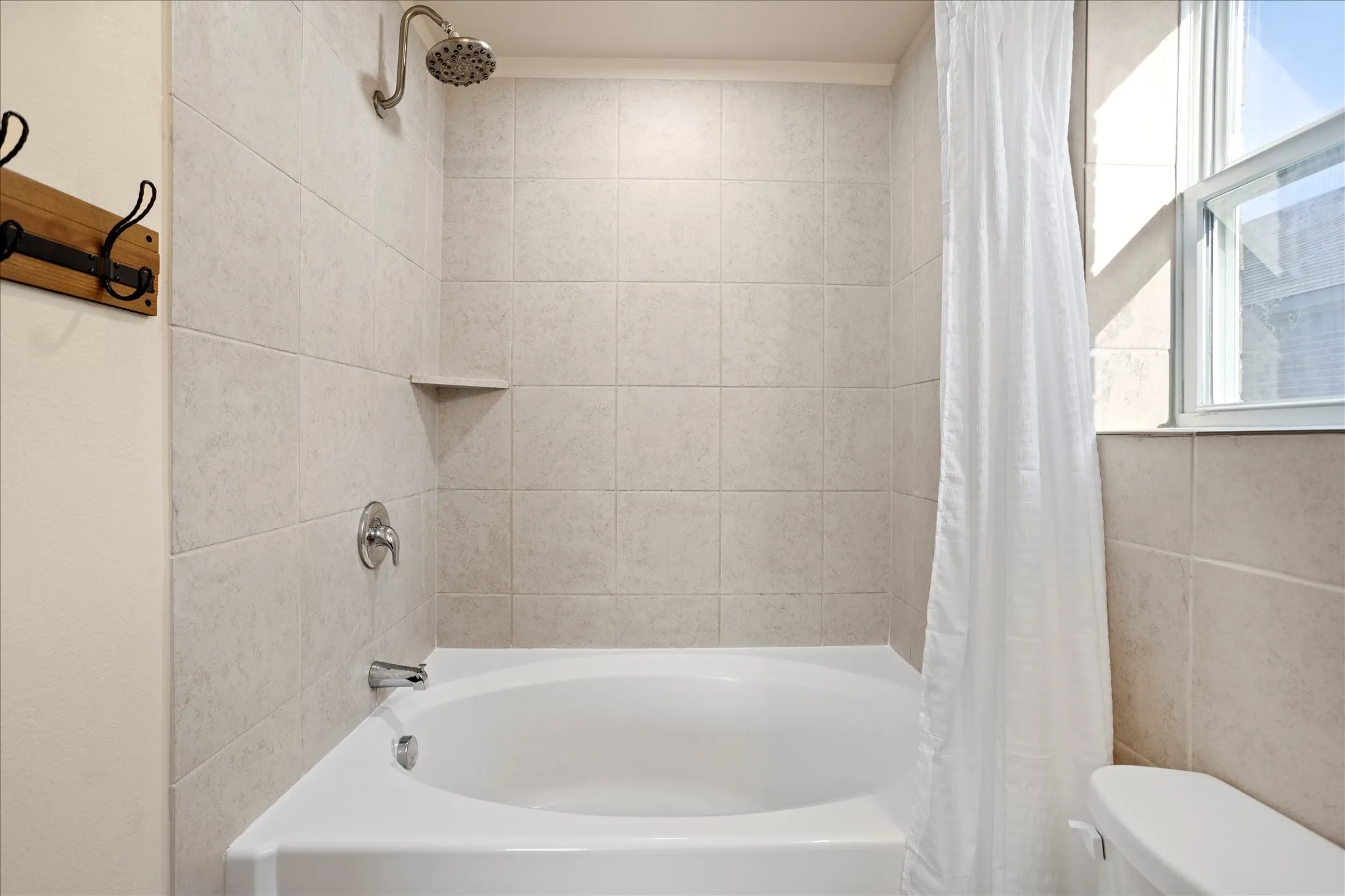 Tub and shower combination with light-toned wall tiling, built-in shelving, a rainfall showerhead, and a window for natural illumination
