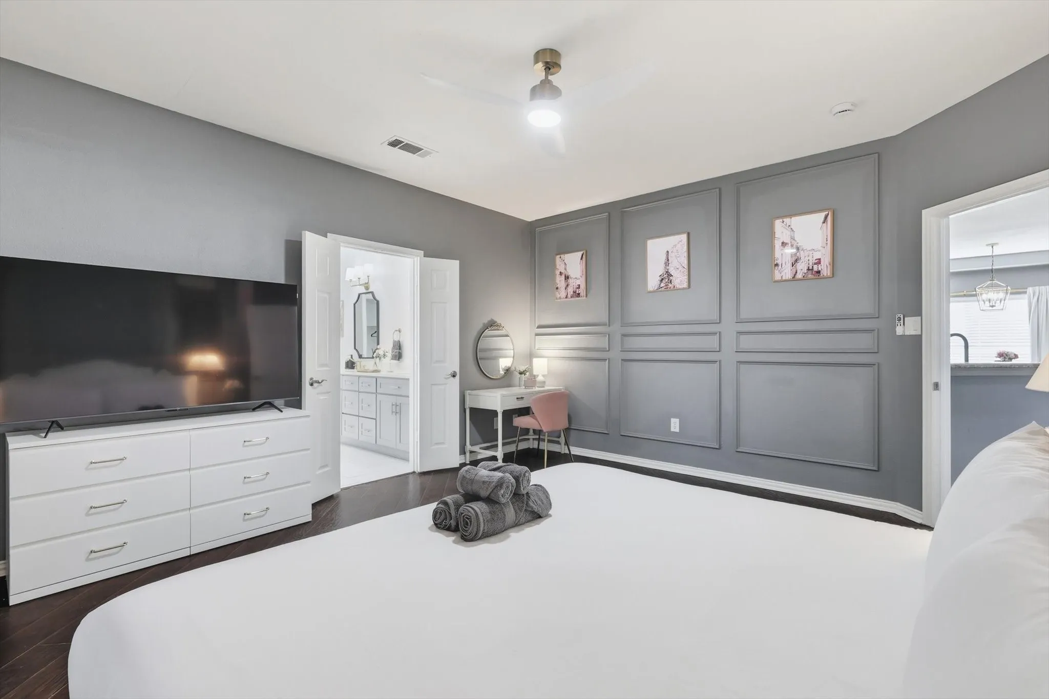 Wood-finish flooring, accent wall with molding, ceiling fan with integrated lighting, dual doors, and an open doorway reveal a vanity with dual sinks