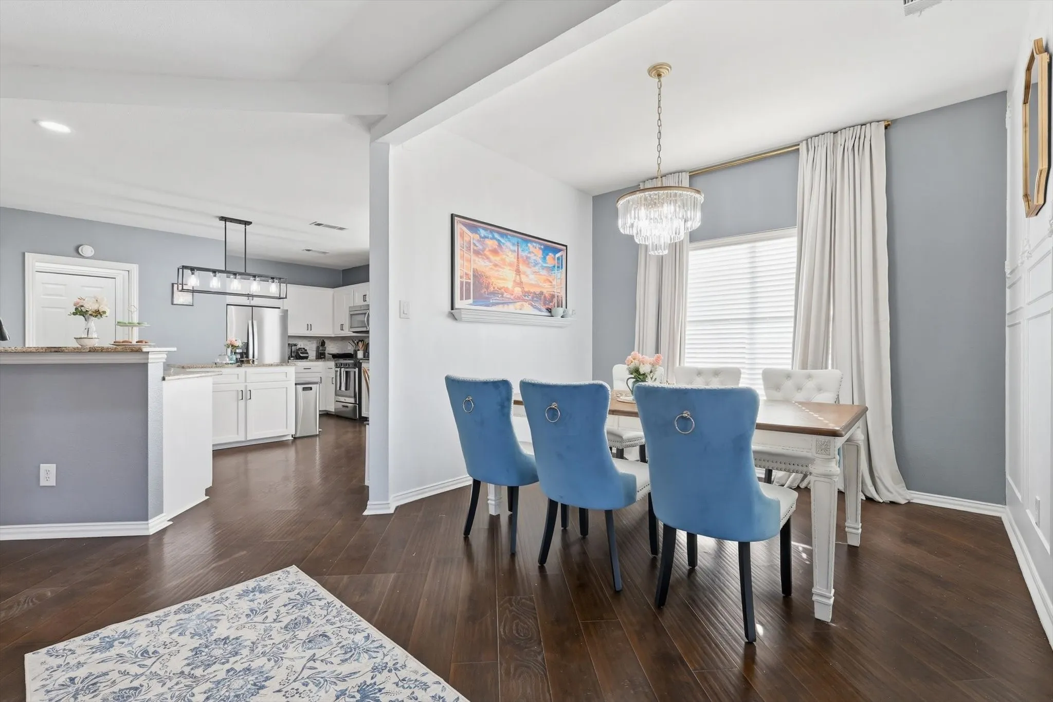 Dark wood-finish flooring throughout, a dining area featuring a crystal chandelier and window with blinds, and a kitchen with white cabinetry and a linear pendant light fixture