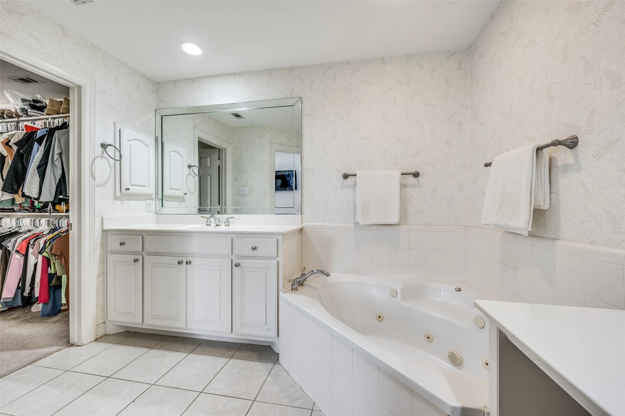 White cabinetry, large vanity mirror, tile flooring, built-in jetted tub, and textured wall coverings