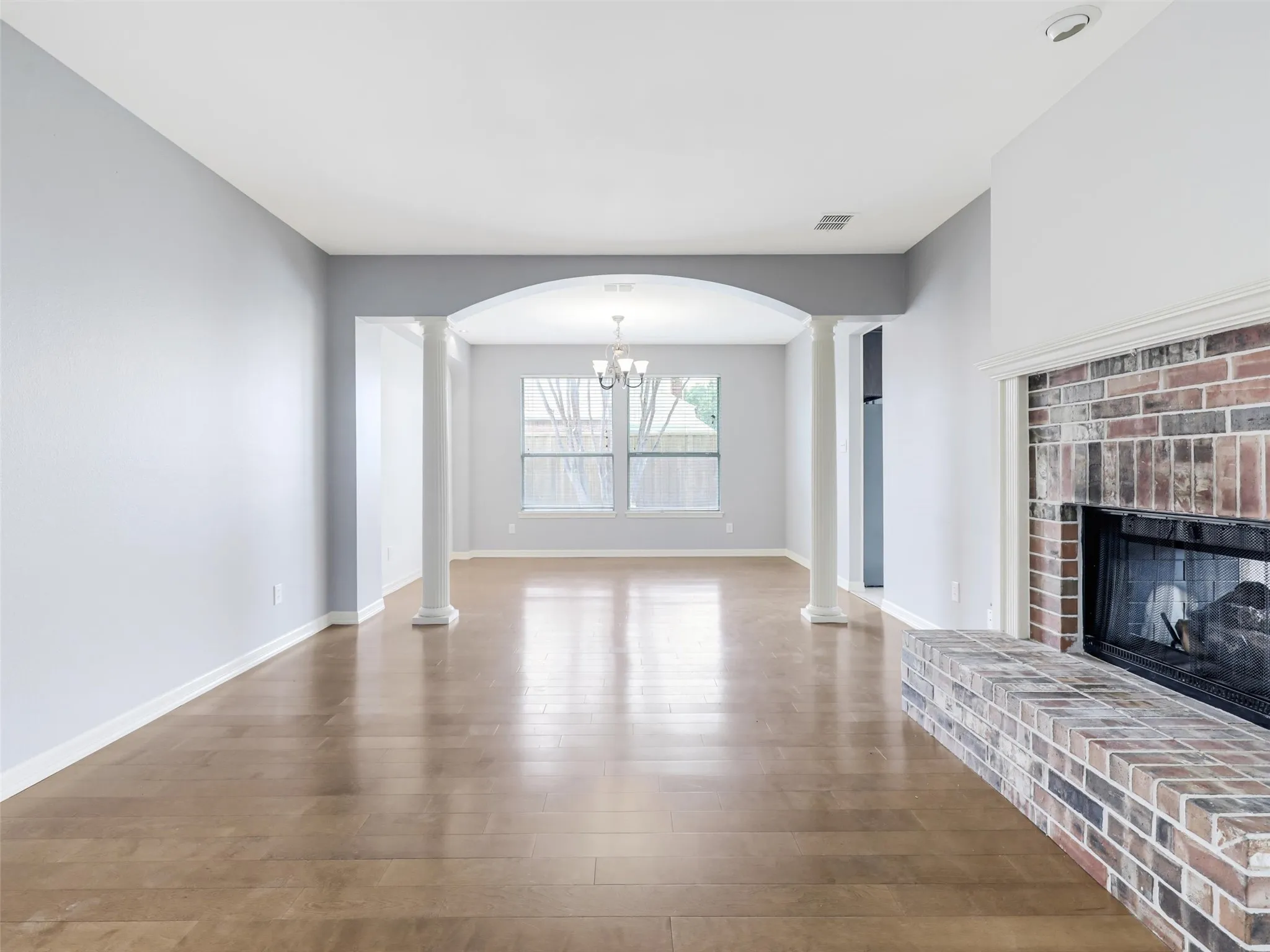 Spacious room with wood-finish flooring, a brick fireplace, and an arched entryway supported by decorative columns