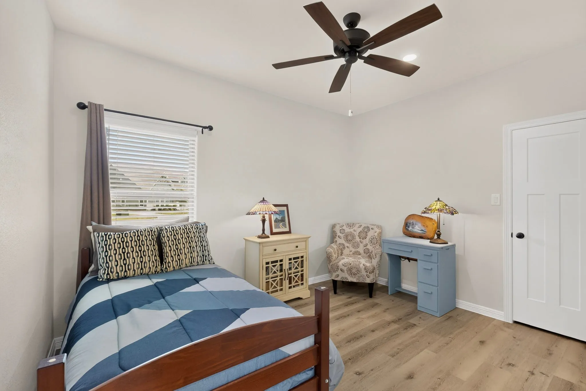 Light-toned walls with wood-finish flooring, a window with blinds, a dark-toned ceiling fan, and a walk in closet