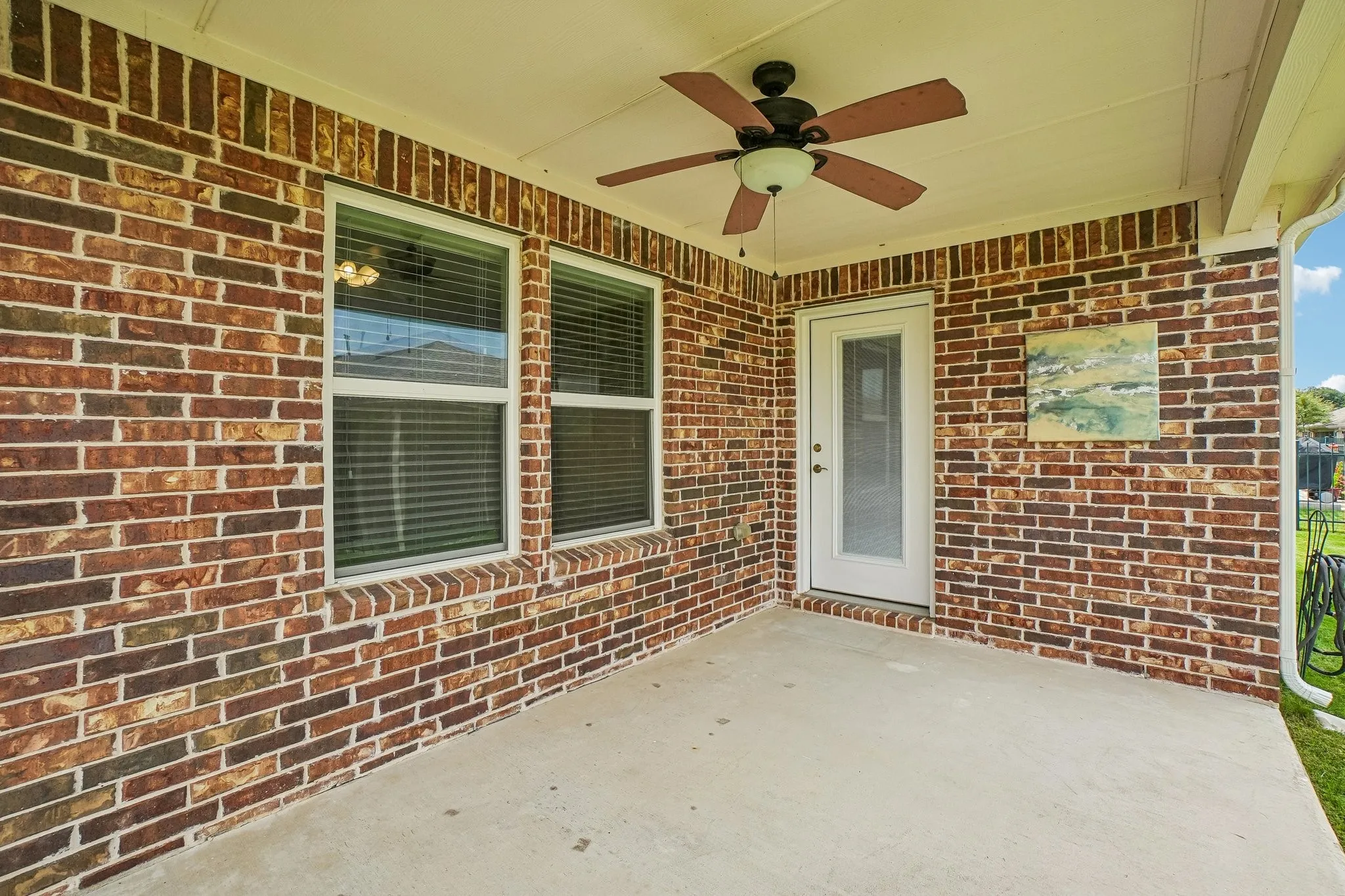 Covered patio featuring brick exterior walls, a ceiling fan, a white exterior door with glass insert, and two windows with blinds