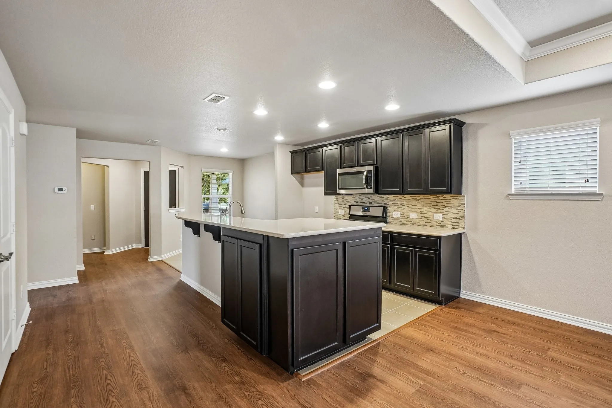 Kitchen island with seating, dark wood-finish cabinetry, stainless steel microwave, patterned tile backsplash, and recessed lighting