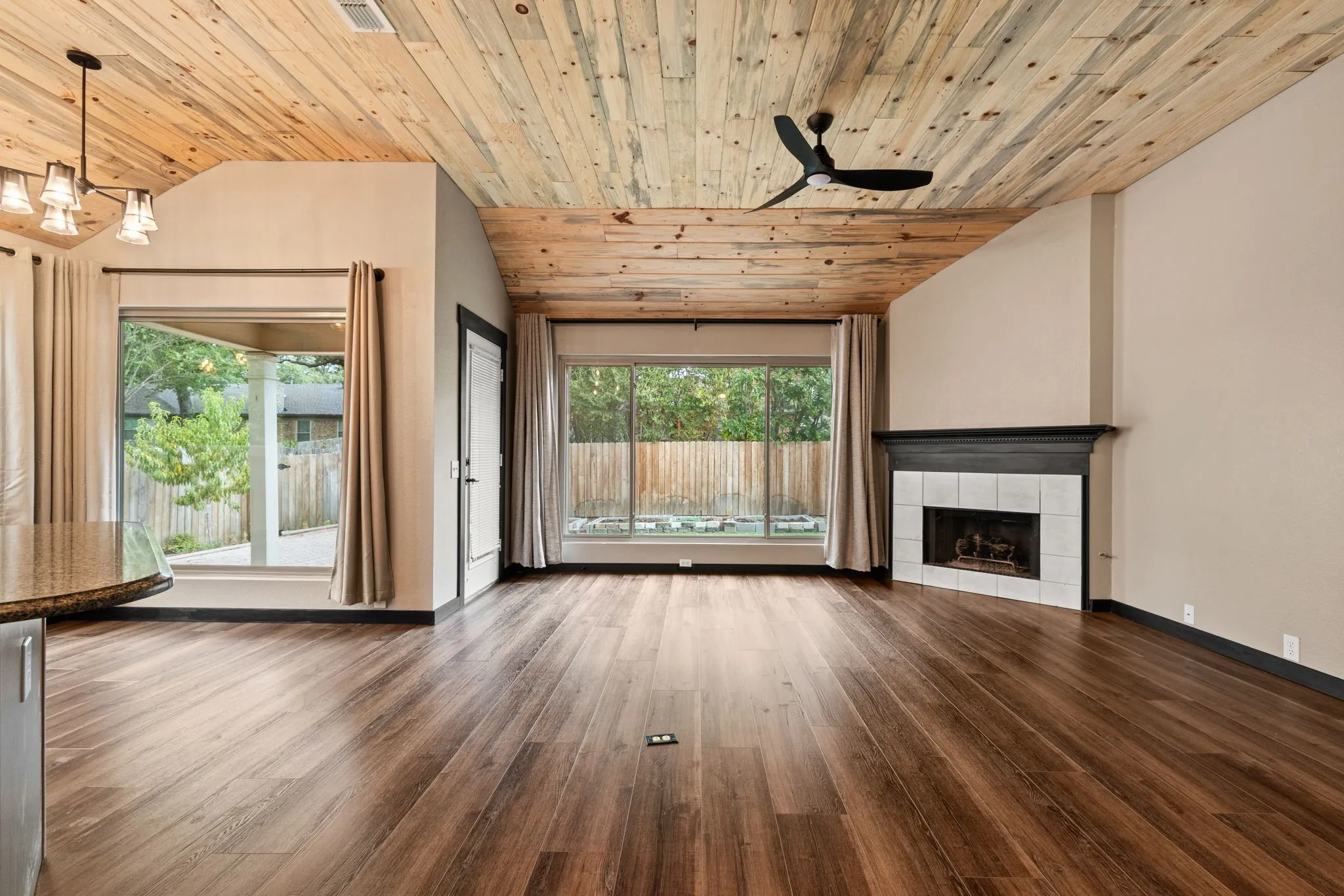 Vaulted wood-finish ceiling, wood-finish flooring, corner fireplace with tiled surround, large windows, and a ceiling fan with integrated lighting