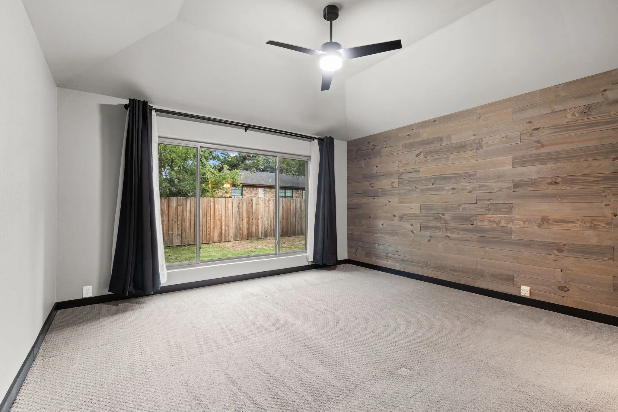 Vaulted ceiling with a ceiling fan, accent wall with wood paneling, light-toned carpet, and a large window