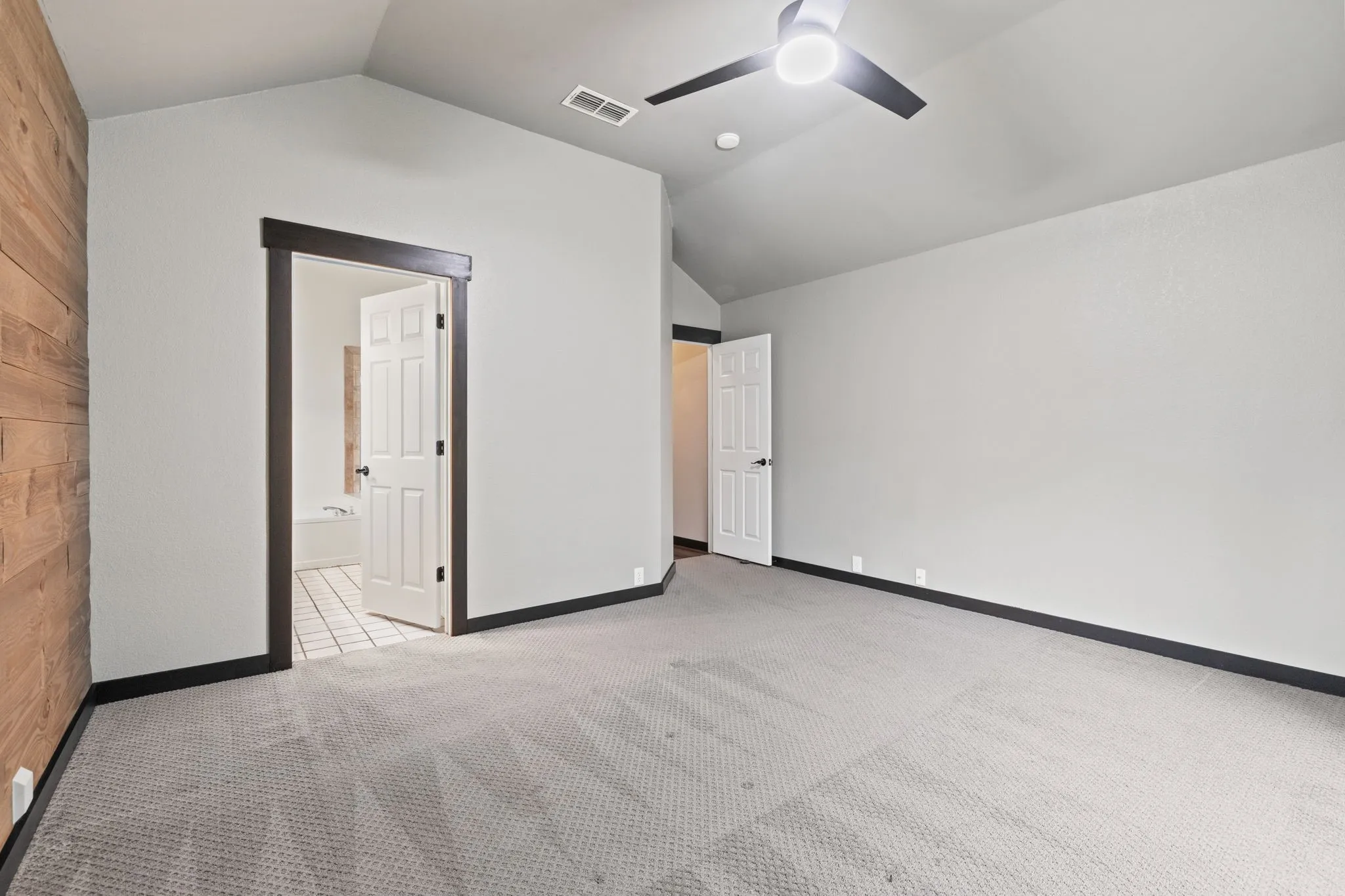 Vaulted ceiling with a ceiling fan, accent wall with wood paneling, light-colored carpeting, dark trim, and a paneled interior door