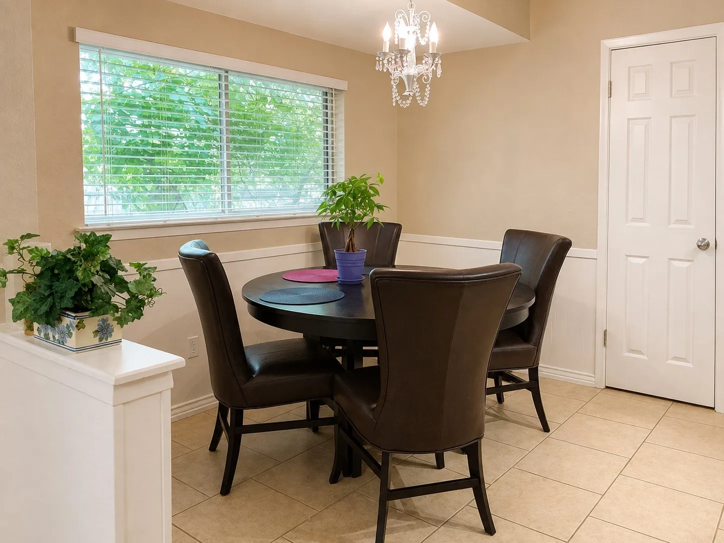 Recessed ceiling with a crystal chandelier, neutral wall tones with wainscoting, large window with blinds, paneled interior door, and tiled flooring