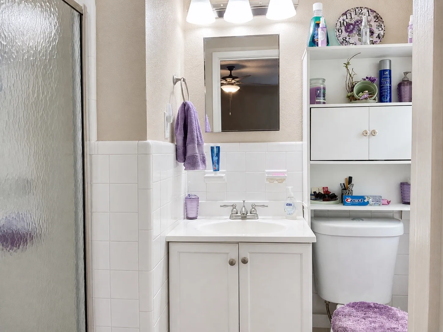 White tile shower enclosure, single vanity with integrated sink, white storage cabinet, wall-mounted mirror, and overhead light fixture