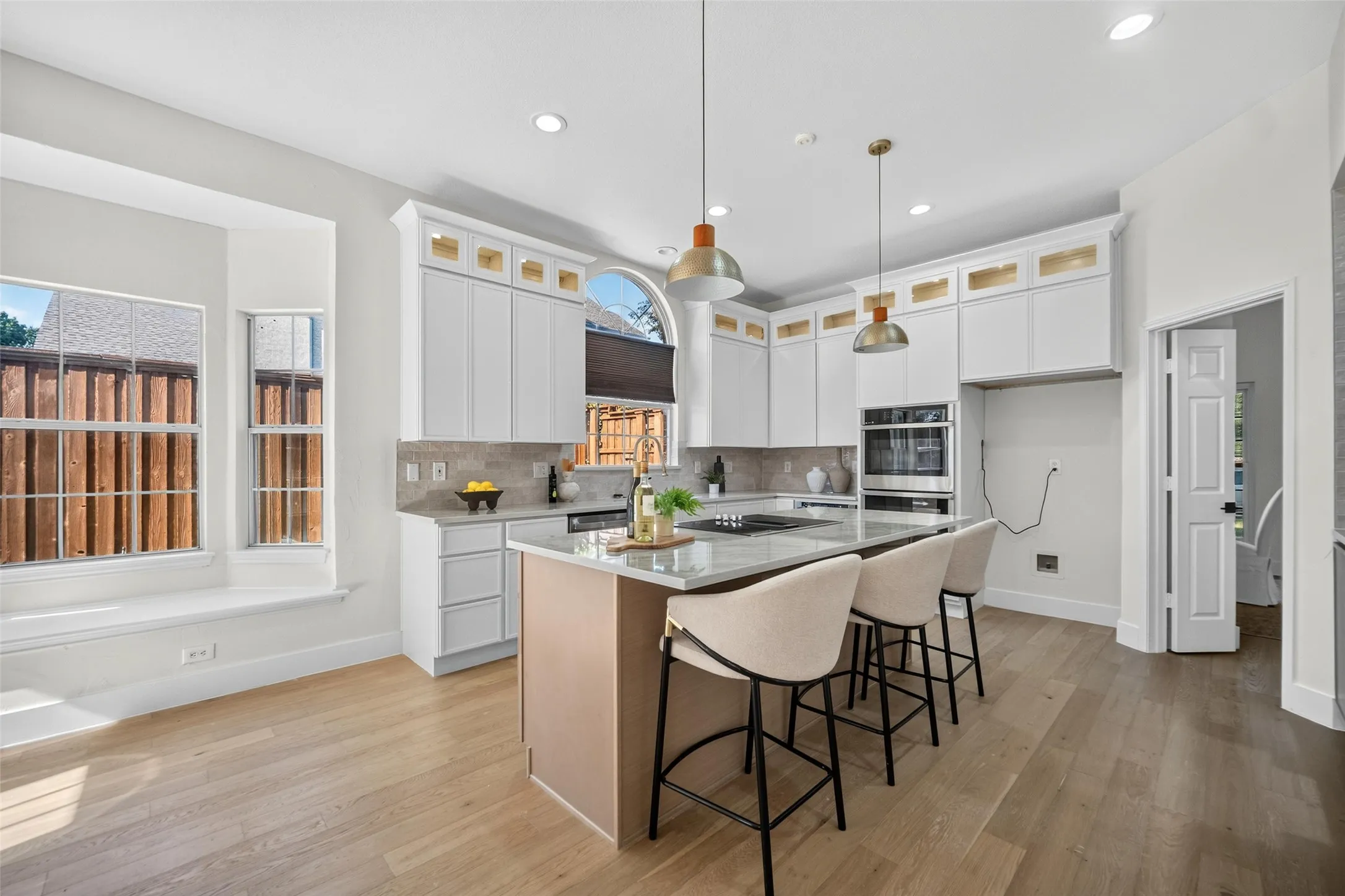 Light wood-finish flooring, white cabinetry with gold-tone accents, an island with countertop seating, a built-in oven, and recessed lighting