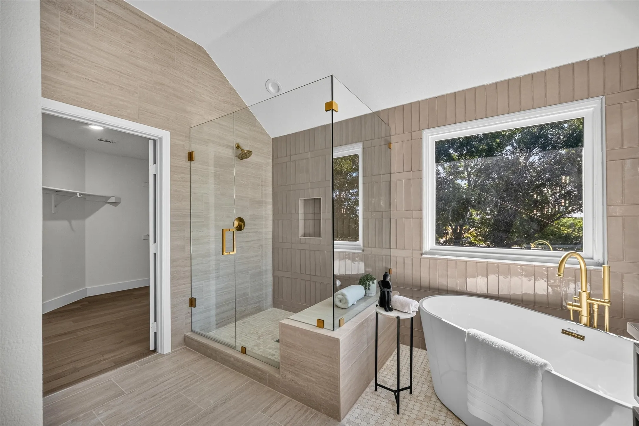 Free-standing soaking tub with brass fixtures, glass-enclosed shower with brass hardware, tiled shower niche, wood-finish flooring, and a walk-in closet