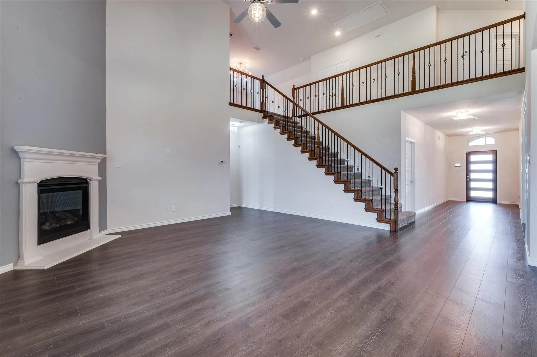Two-story entry, wood-finish flooring, staircase with dark wood handrails and wrought iron balusters, fireplace with white mantel, and ceiling fan with integrated lighting