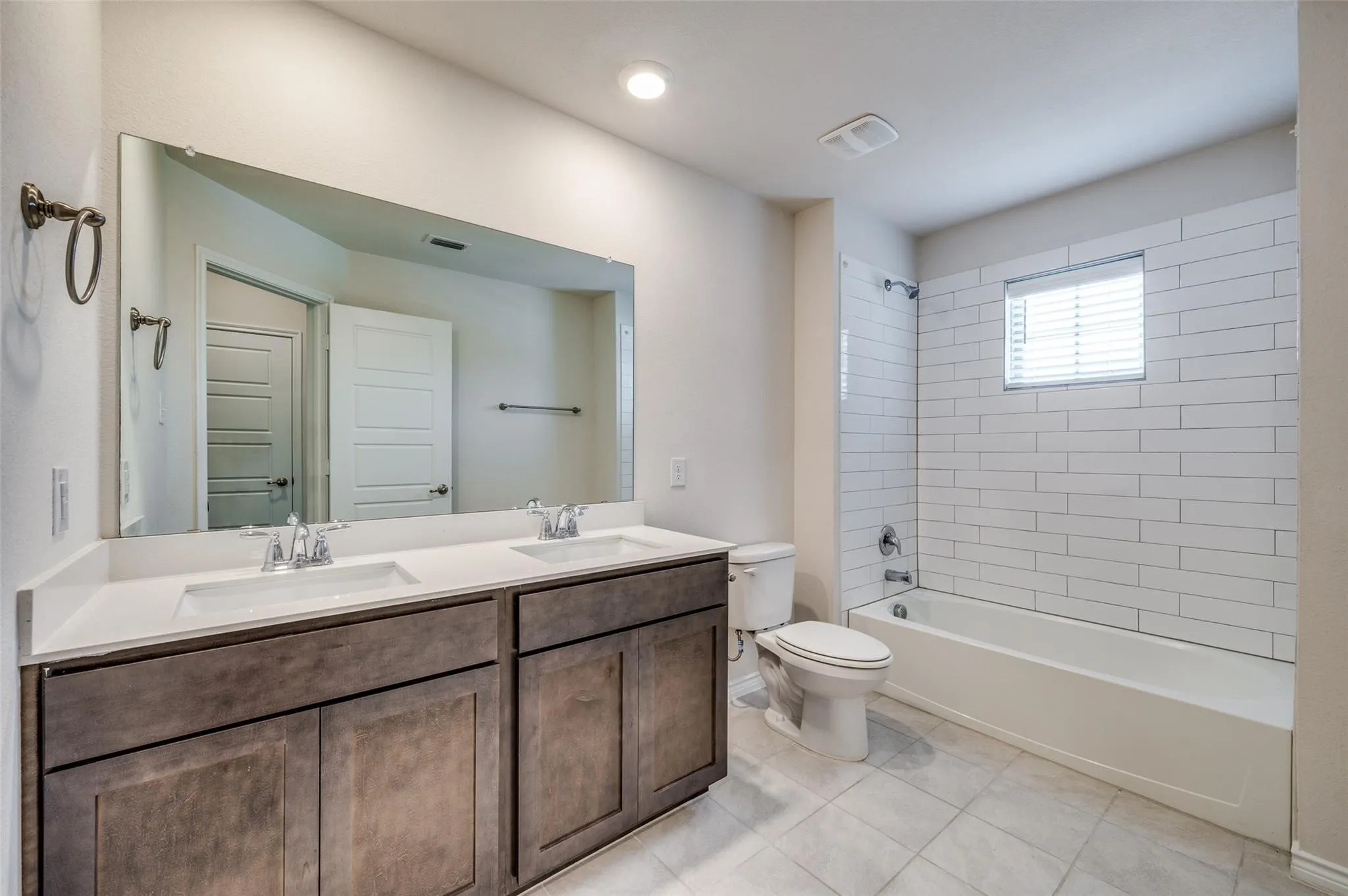 Dual vanity with integrated sinks and wood-finish cabinetry, a large vanity mirror, a bathtub with a subway tile surround, and tile flooring