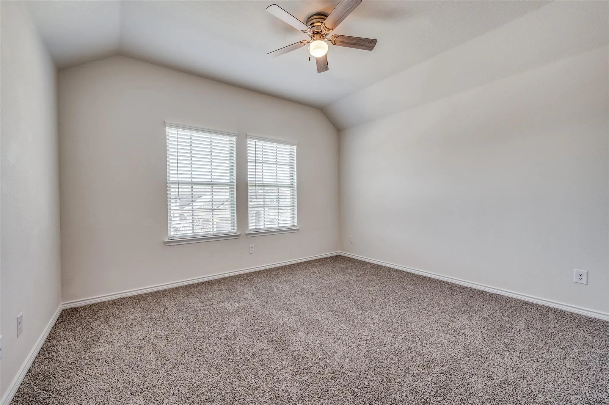 Vaulted ceiling, ceiling fan with integrated lighting, two windows with blinds, neutral wall paint, and textured carpeting