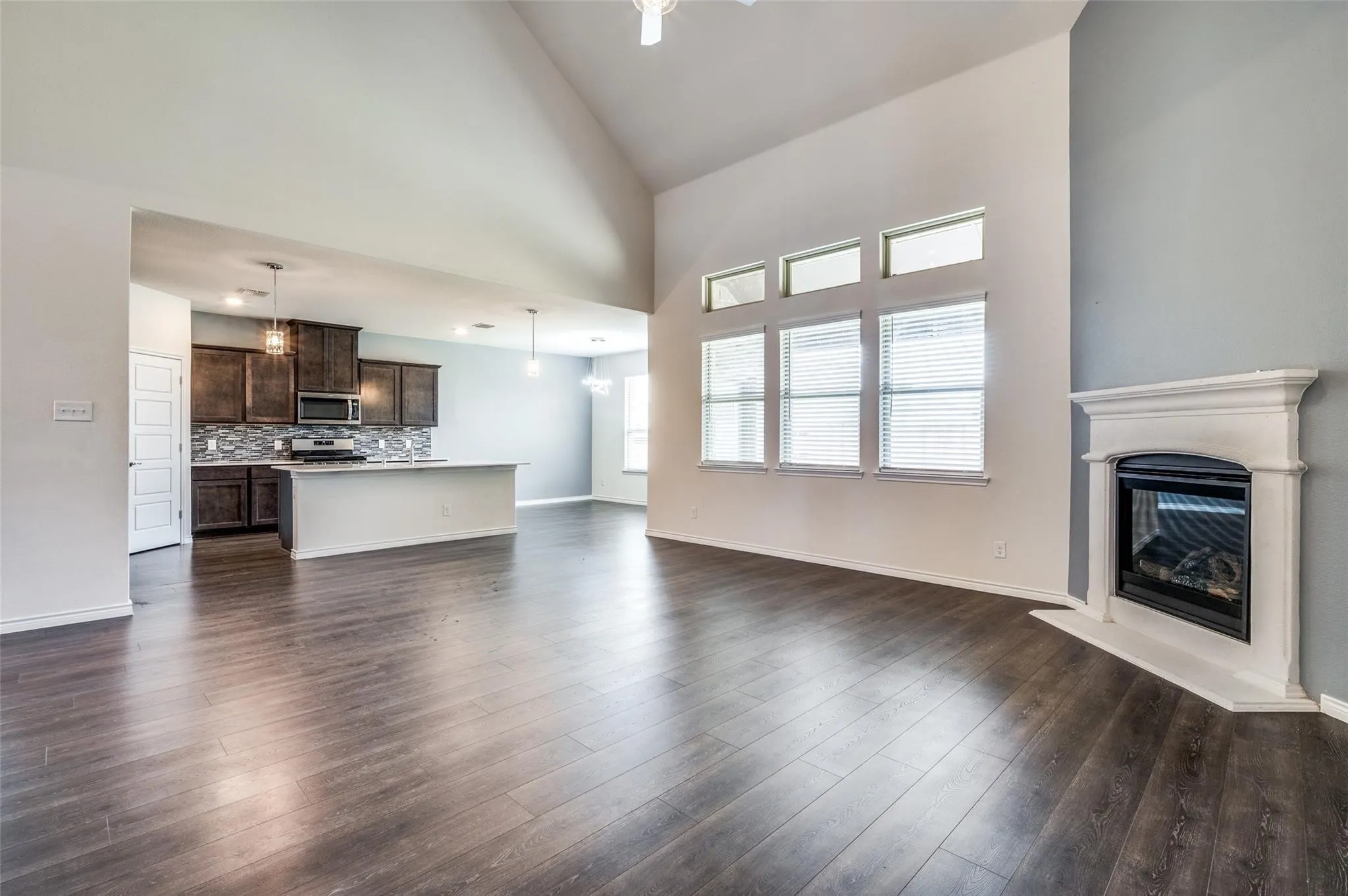 Vaulted ceilings, wood-finish flooring, a corner fireplace with a white mantel, kitchen island with pendant lighting, and dark wood cabinetry