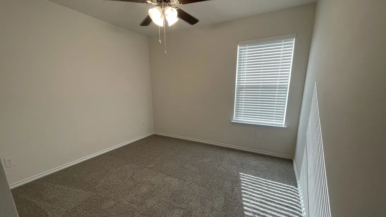 Neutral wall color, textured carpeting, window with horizontal blinds, and a ceiling fan with integrated lighting