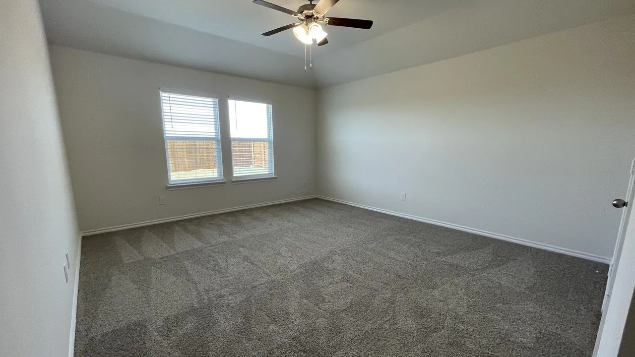 Spacious room with neutral-toned carpet flooring, two large windows with blinds, a ceiling fan with integrated lighting, and light-colored walls