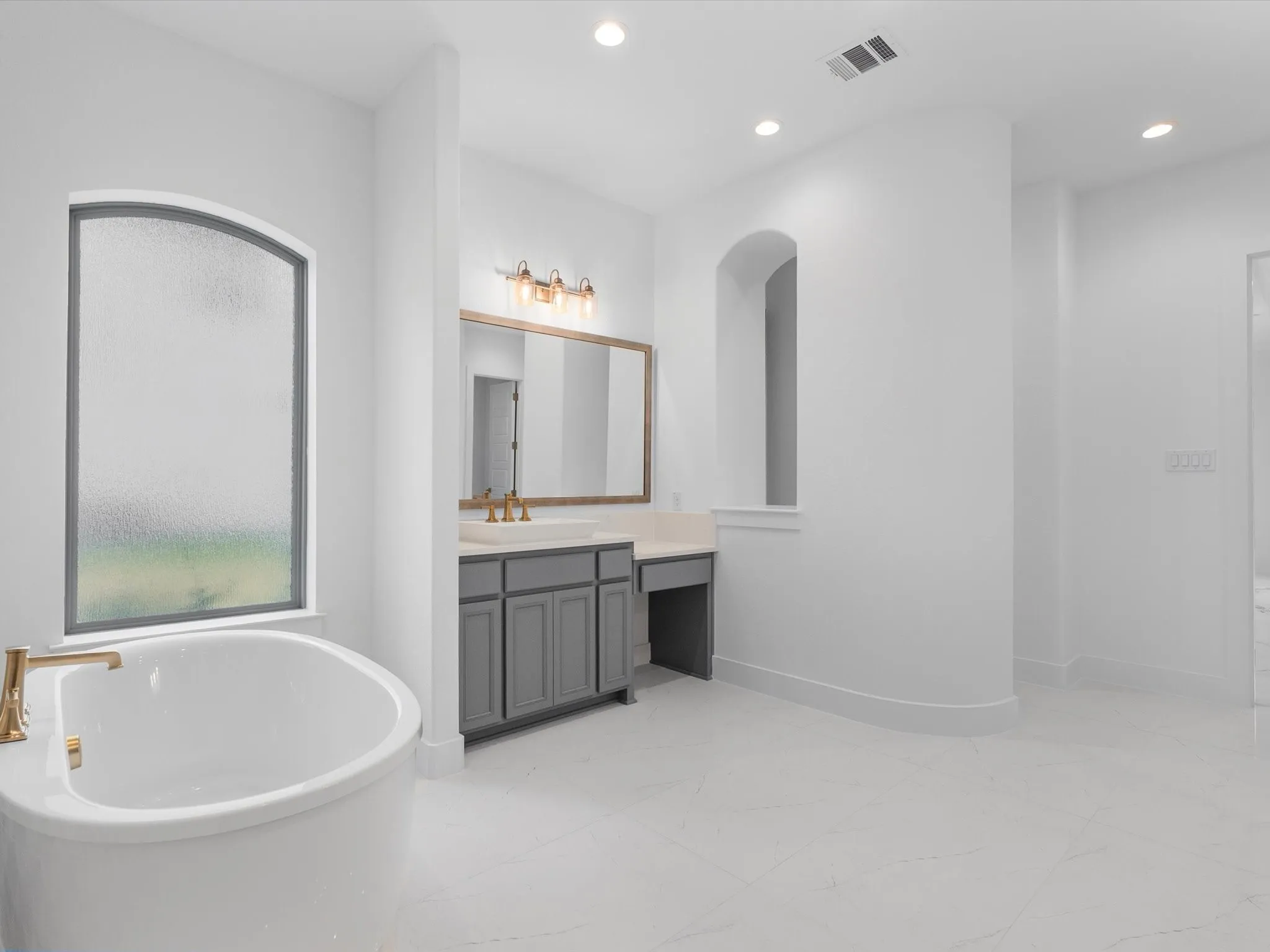 Spa-inspired primary bathroom featuring a freestanding soaking tub with brass fixtures, dual-sink vanity with gray cabinetry and wood-finish framing, quartz countertops, brass-finished lighting, and arched architectural detailing