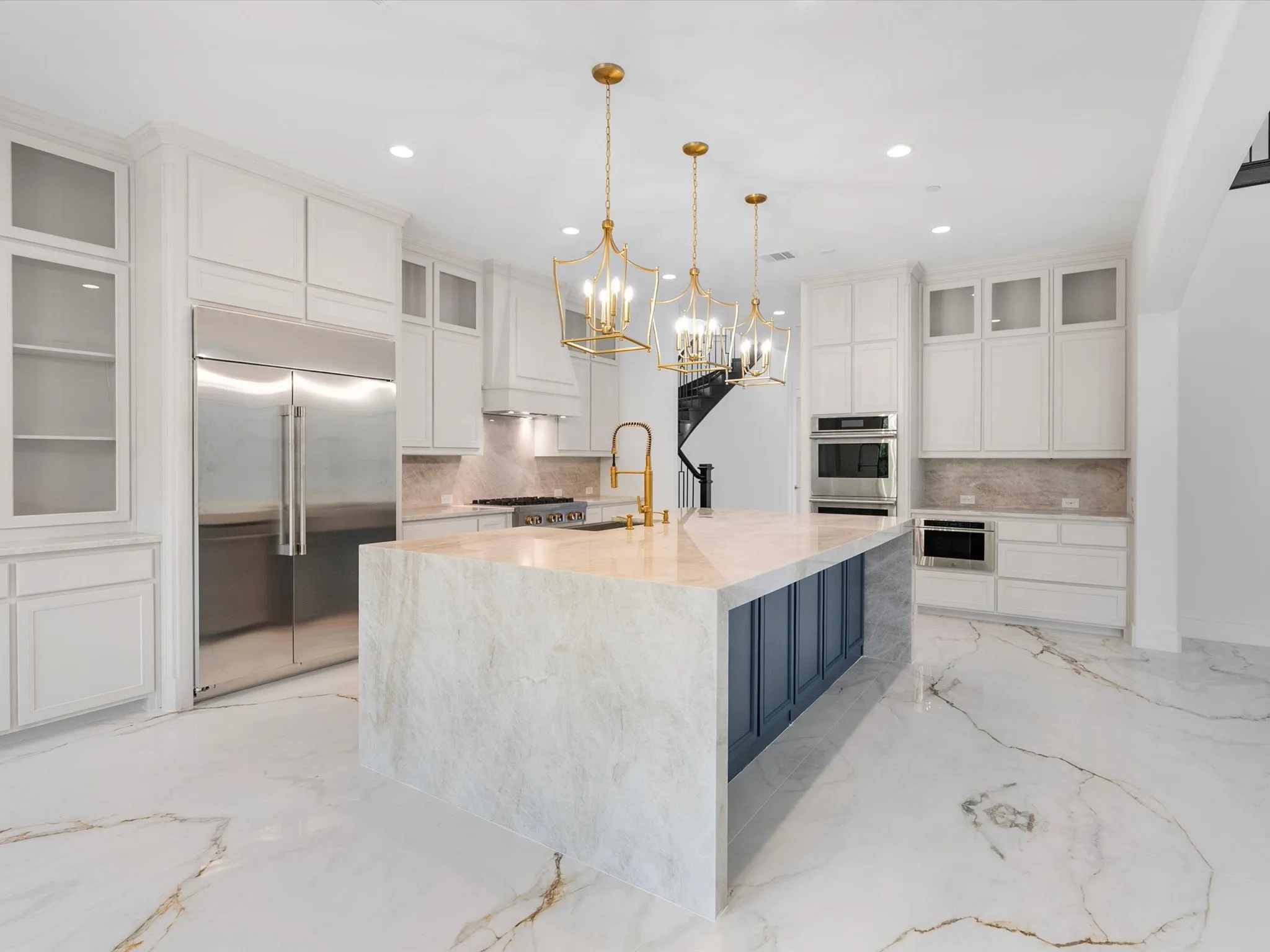 Expansive kitchen island with waterfall countertop, stainless steel appliances, white cabinetry with glass-front accents, and gold-finish pendant lighting