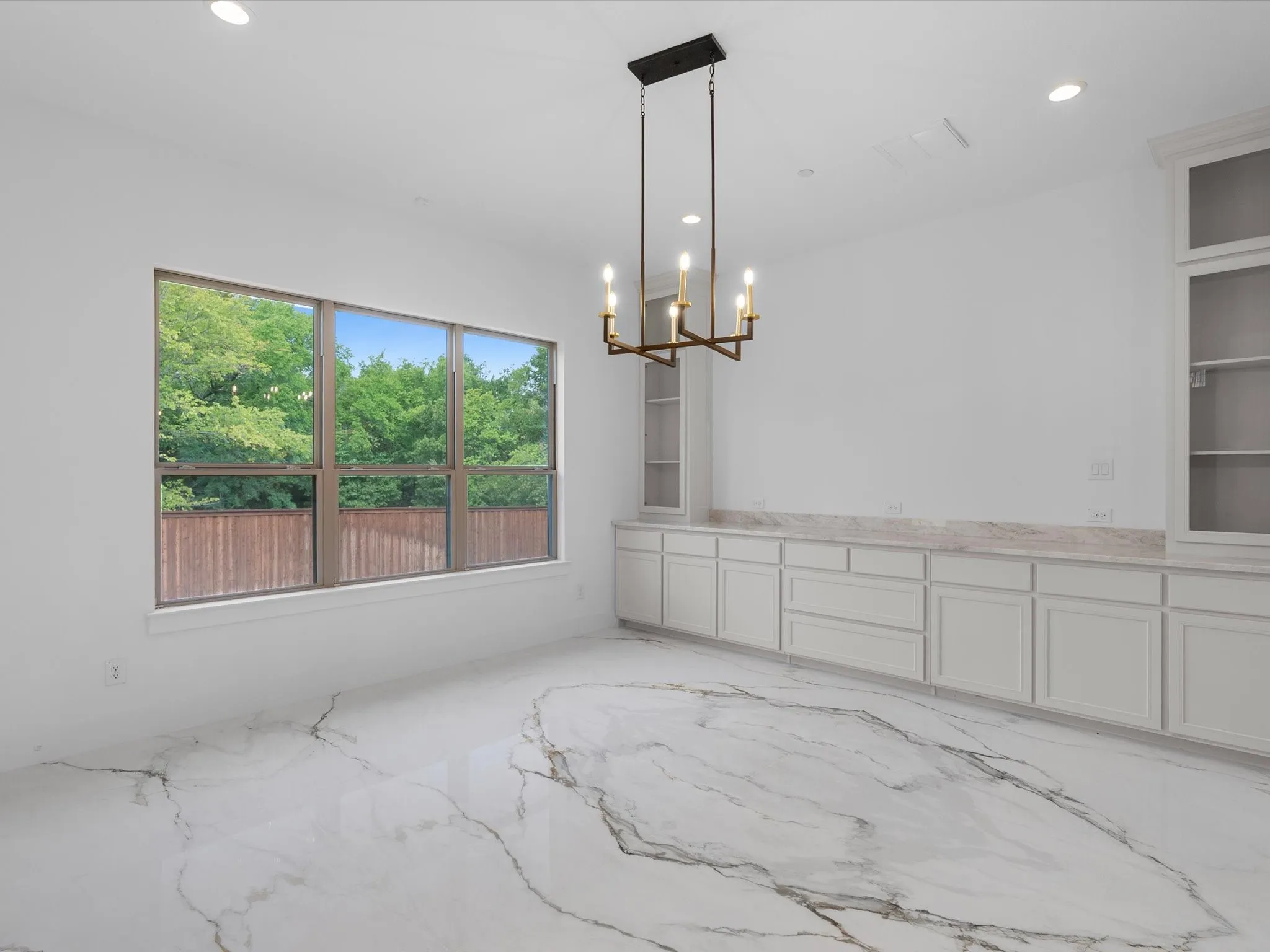 Spacious room featuring large format marble-look tile flooring, white built-in cabinetry with stone countertops, a contemporary chandelier, and a large window overlooking a wooden fence and mature trees