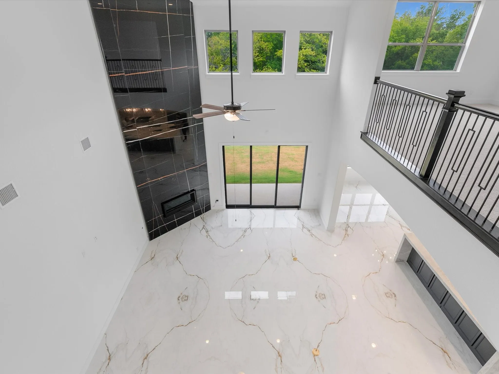 Two-story space featuring a floor-to-ceiling black marbled accent wall, an electric fireplace, a ceiling fan, and large format polished tile flooring