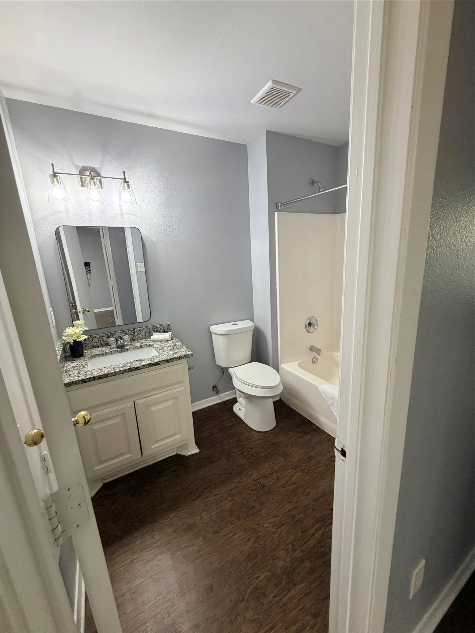 Bathroom featuring a single vanity with granite-style countertop, white cabinetry, rectangular mirror, wood-finish flooring, and a shower-tub combination