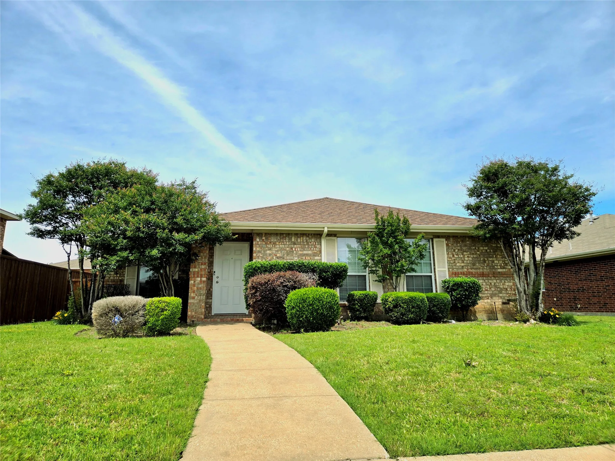 Brick exterior with a brown shingle roof, established landscaping, and a concrete pathway
