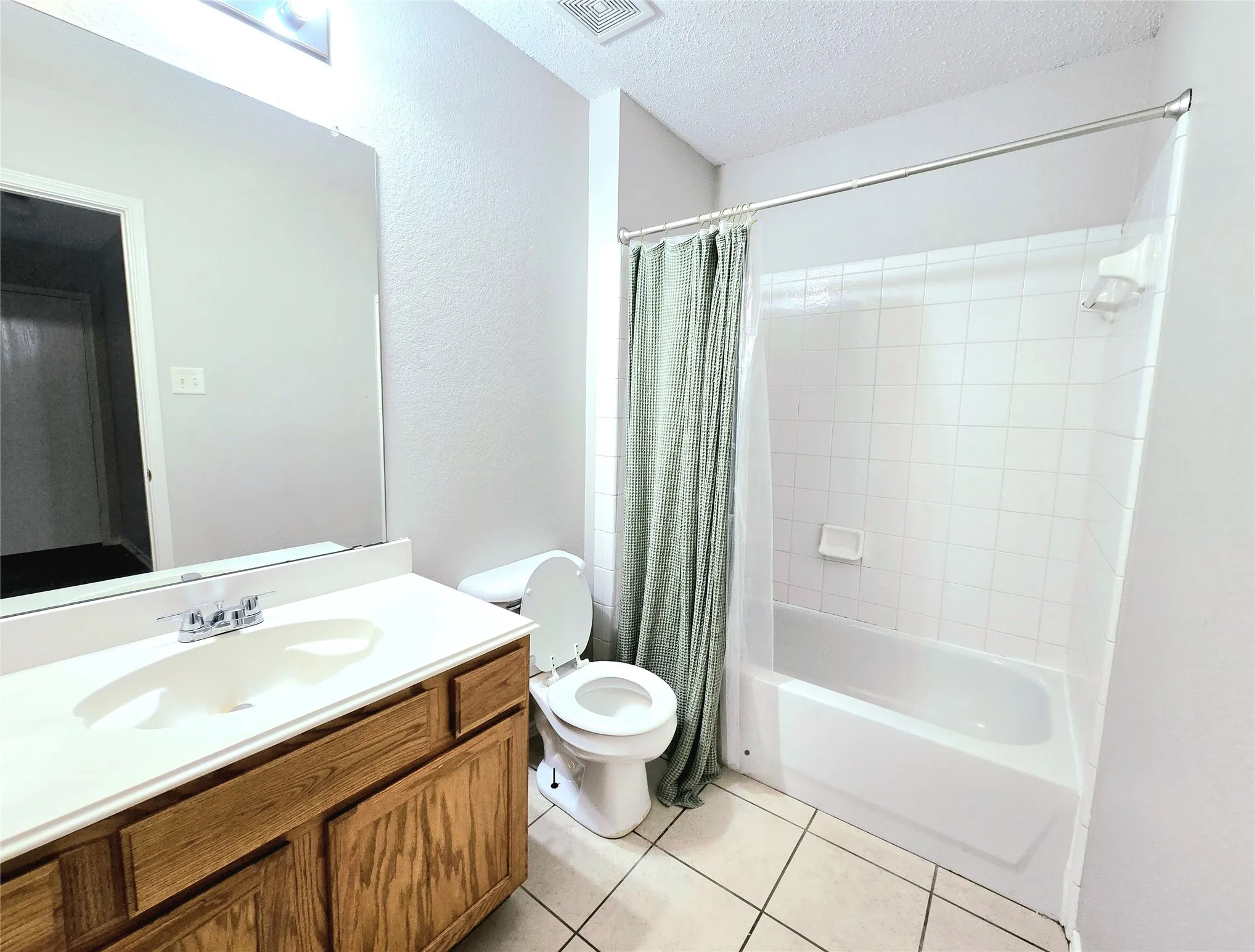Bright bathroom featuring a wood-finish vanity with dual sinks, large framed mirror, ceramic tile flooring, and a combined shower-tub enclosure with tile surround and coordinating shower curtain rod