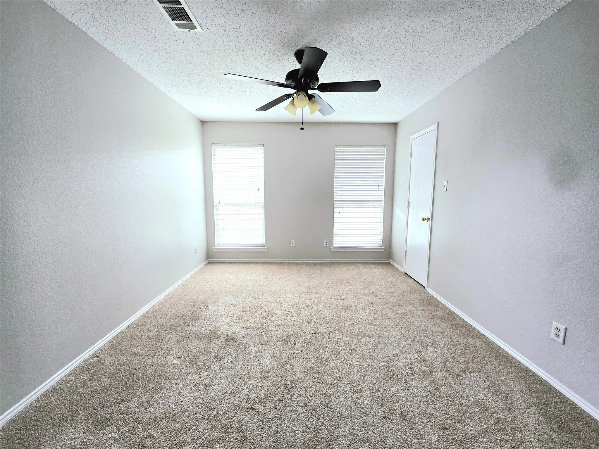 Light gray wall paint, tan carpeting, two windows with blinds, a ceiling fan with light, and a white door with a brass knob