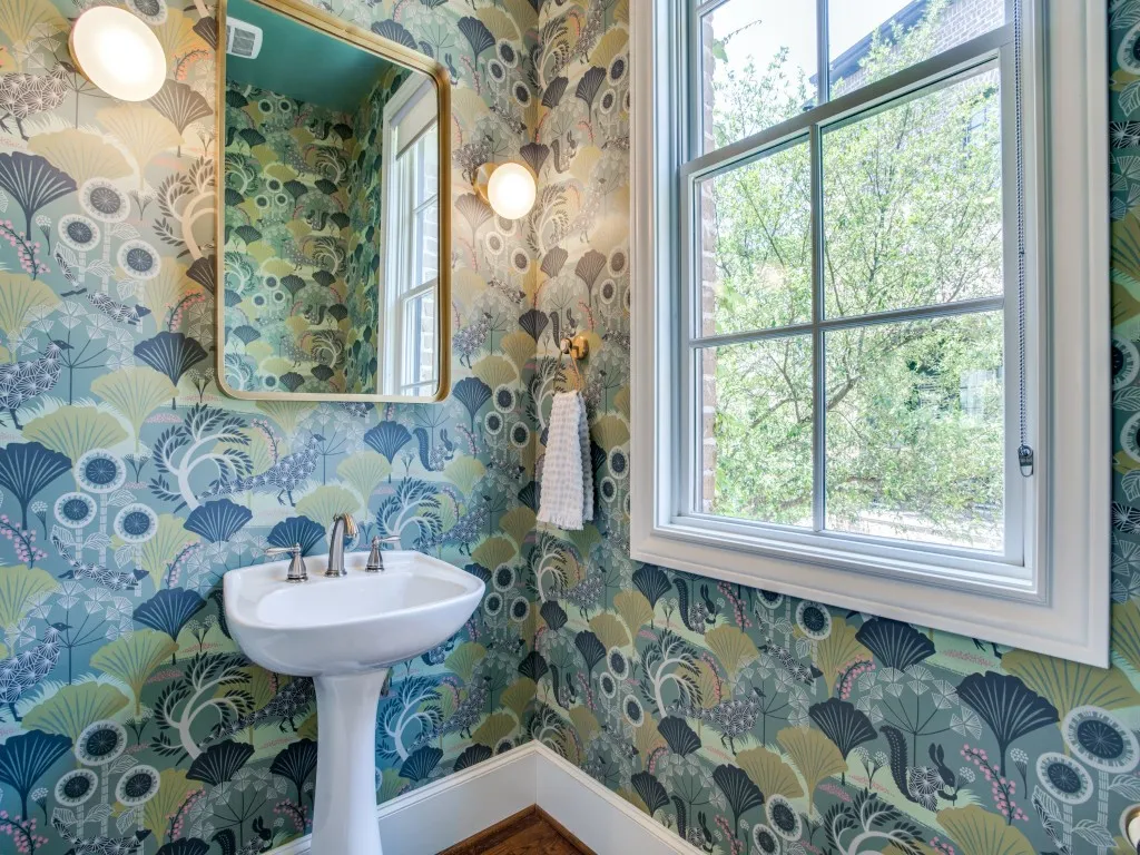 Pedestal sink with a polished chrome faucet, decorative wall sconces, gold-framed mirror, and patterned wallpaper