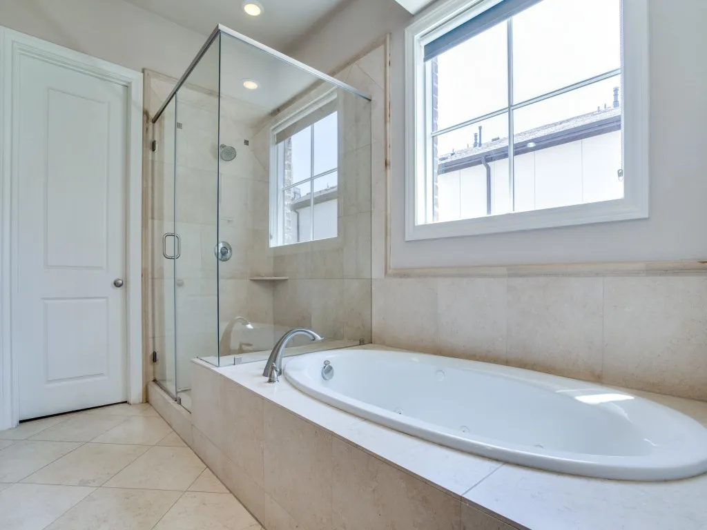 Tiled shower enclosure with a clear glass door, recessed lighting, a built-in tub with a deck-mounted faucet, and a large window