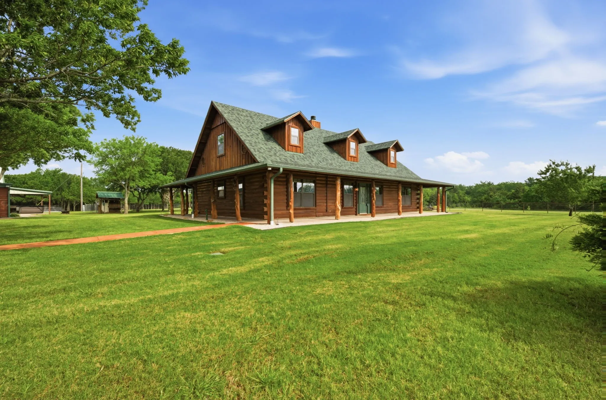 Log cabin exterior with a green shingle roof, multiple dormer windows, and a full-length covered porch featuring natural wood posts
