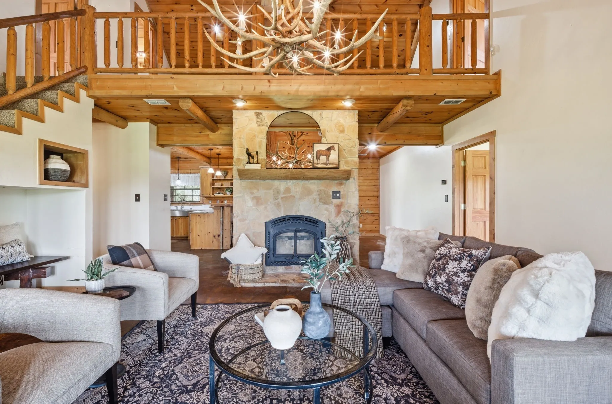 Stone fireplace with hearth, wood-beamed ceiling, wood-finish flooring, staircase with wood railing, and an upper-level wood-railed landing