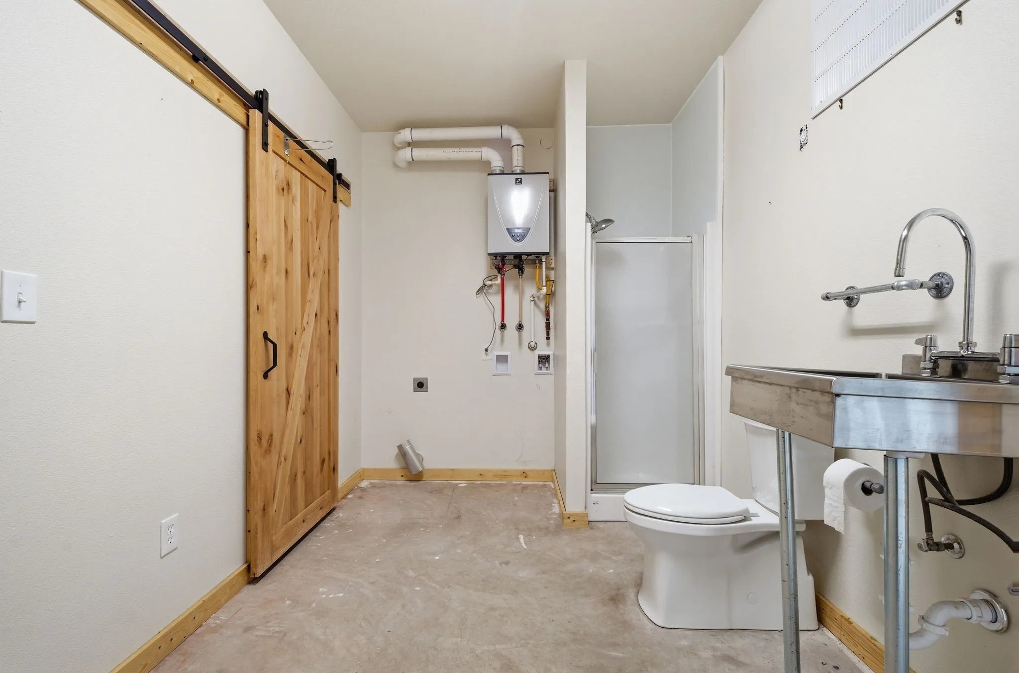 Barn door on a track system, compact shower enclosure, white toilet, wall-mounted water heater, and stainless steel utility sink