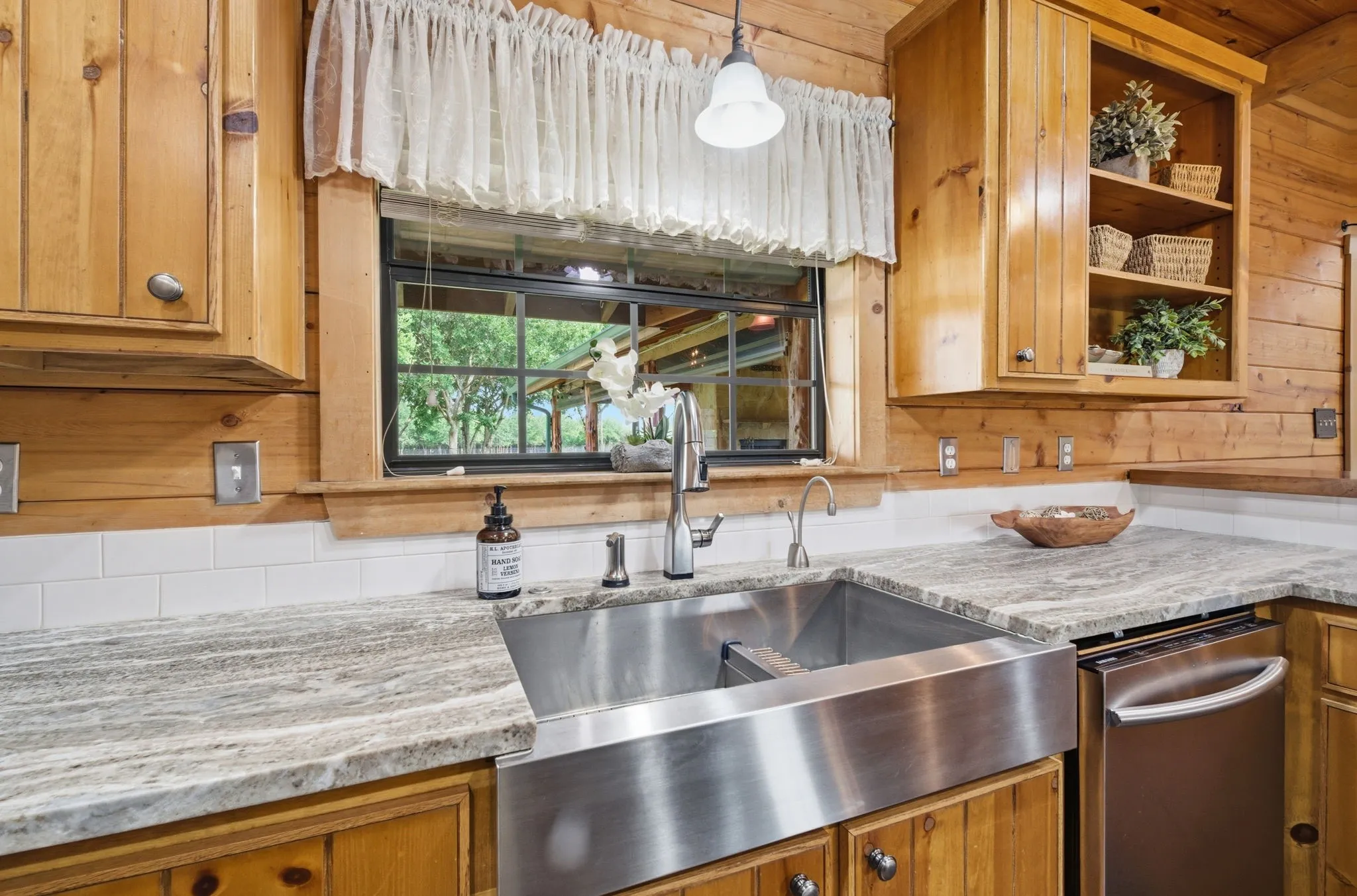 Stainless steel apron-front sink, light-toned countertop, white subway tile backsplash, natural wood cabinetry, and stainless steel dishwasher